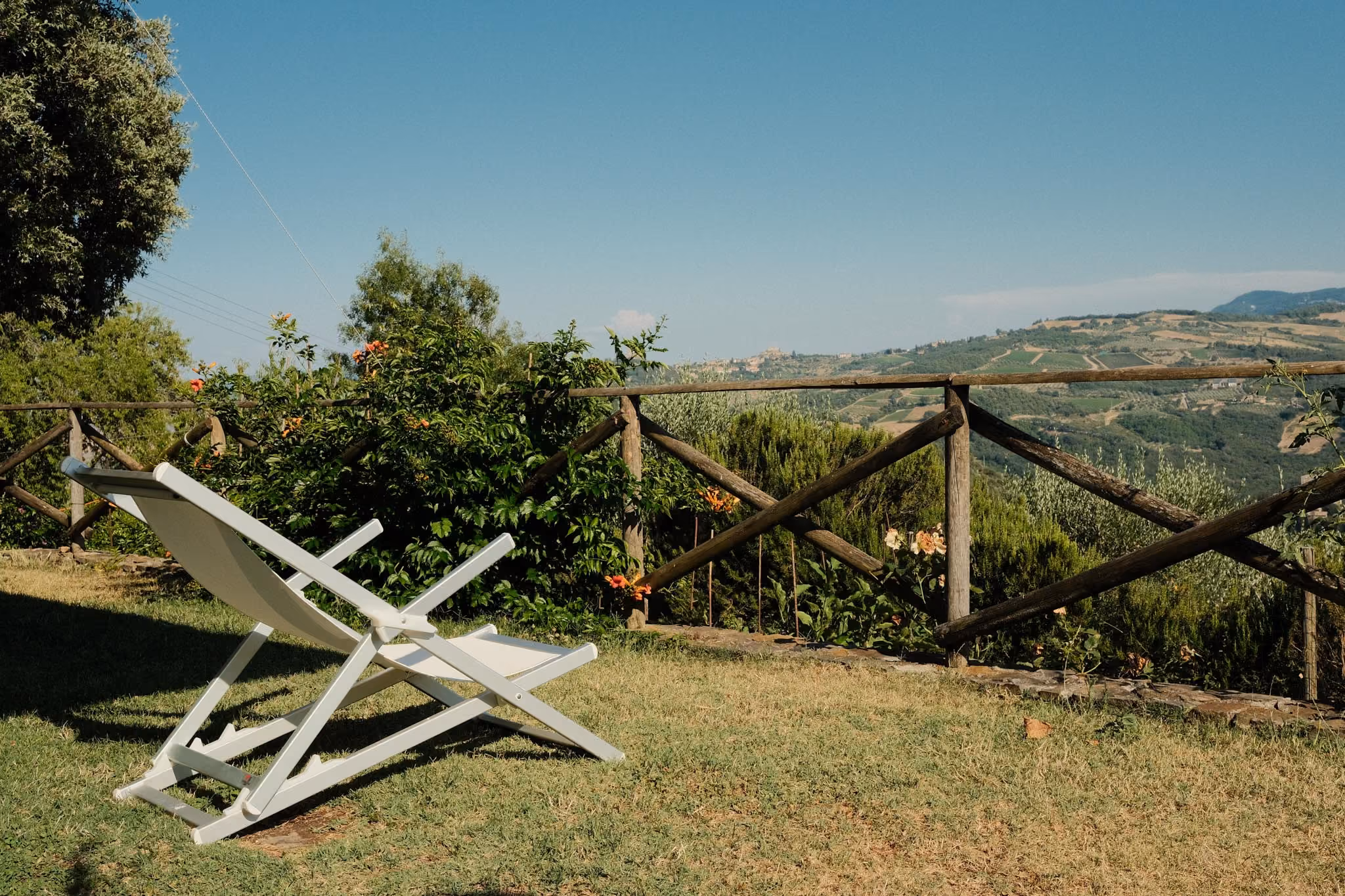 Ein weißer Liegestuhl auf einer Wiese mit Blick auf eine hölzerne Gartenzaun, dahinter Hügel und bewaldete Landschaft unter blauem Himmel.