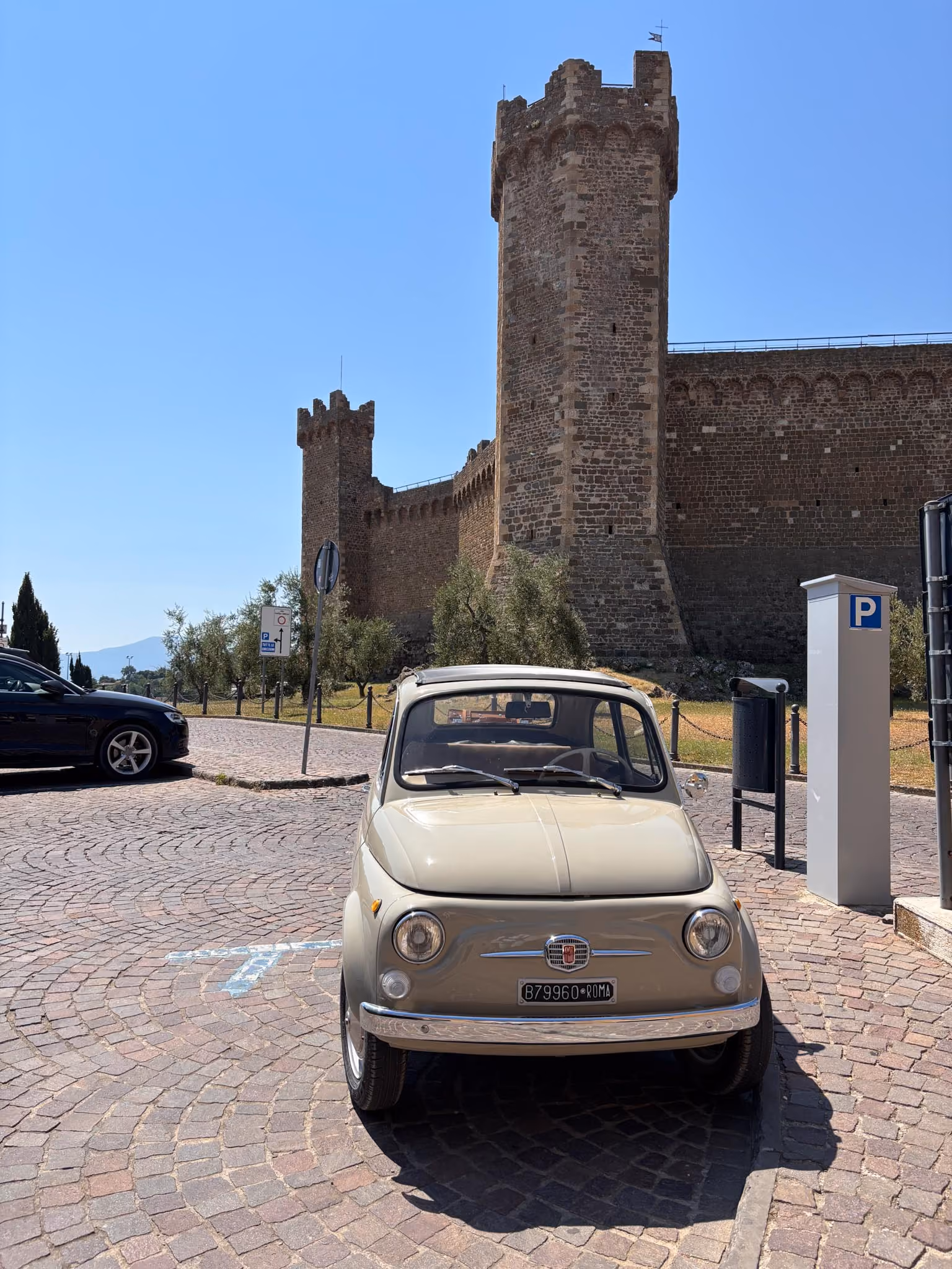 Beiger Fiat 500 Oldtimer geparkt auf einem Kopfsteinpflasterplatz vor einer alten mittelalterlichen Steinburg mit hohen Türmen unter einem klaren blauen Himmel.