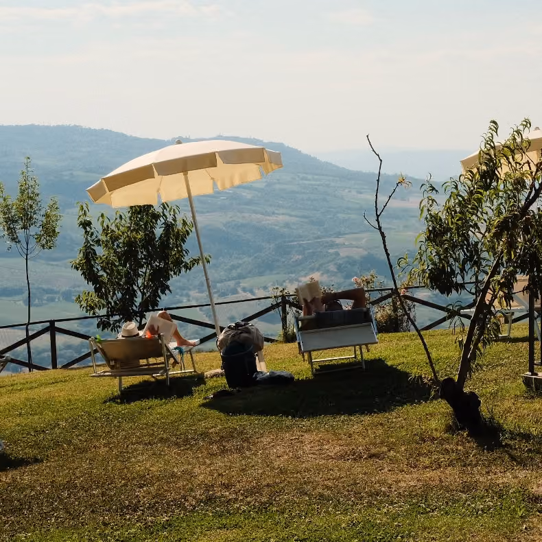 Zwei Personen liegen auf Liegestühlen unter Sonnenschirmen auf einer Wiese mit Blick auf hügelige Landschaft in der Ferne.
