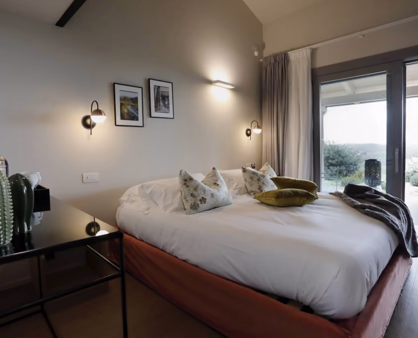 Gemütliches Schlafzimmer mit großem Bett, weißen Bettwäsche und gemusterten Kissen, zwei Wandleuchten, zwei gerahmten Bildern an der Wand, schwarzem Nachttisch mit Dekoration und großen Fenstern mit Blick ins Freie.