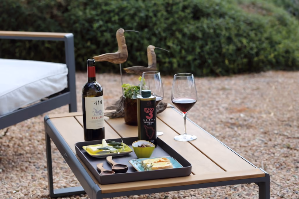 Ein Gartentisch mit zwei hölzernen Vogelstatuen, einer Flasche Rotwein, einem Glas Rotwein, einem Ölbehälter und einem Tablett mit kleinen Schälchen und Brot, neben einem gepolsterten Sitzmöbel im Freien auf Kiesboden.