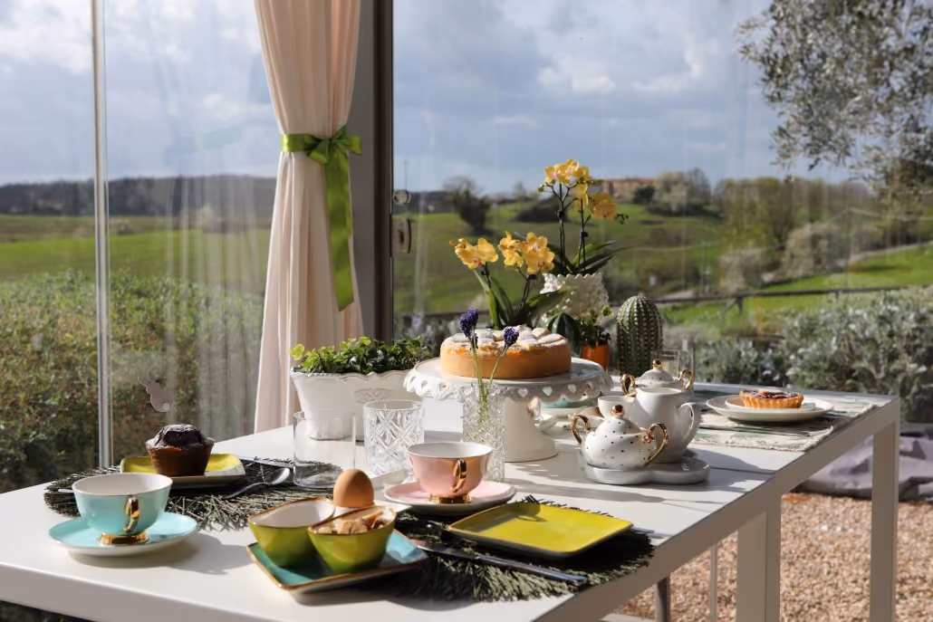 Tisch mit gedecktem Frühstück mit Kuchen, Tassen, Torten und Blumen vor großer Fensterfront mit Blick auf grüne Landschaft bei bewölktem Himmel.