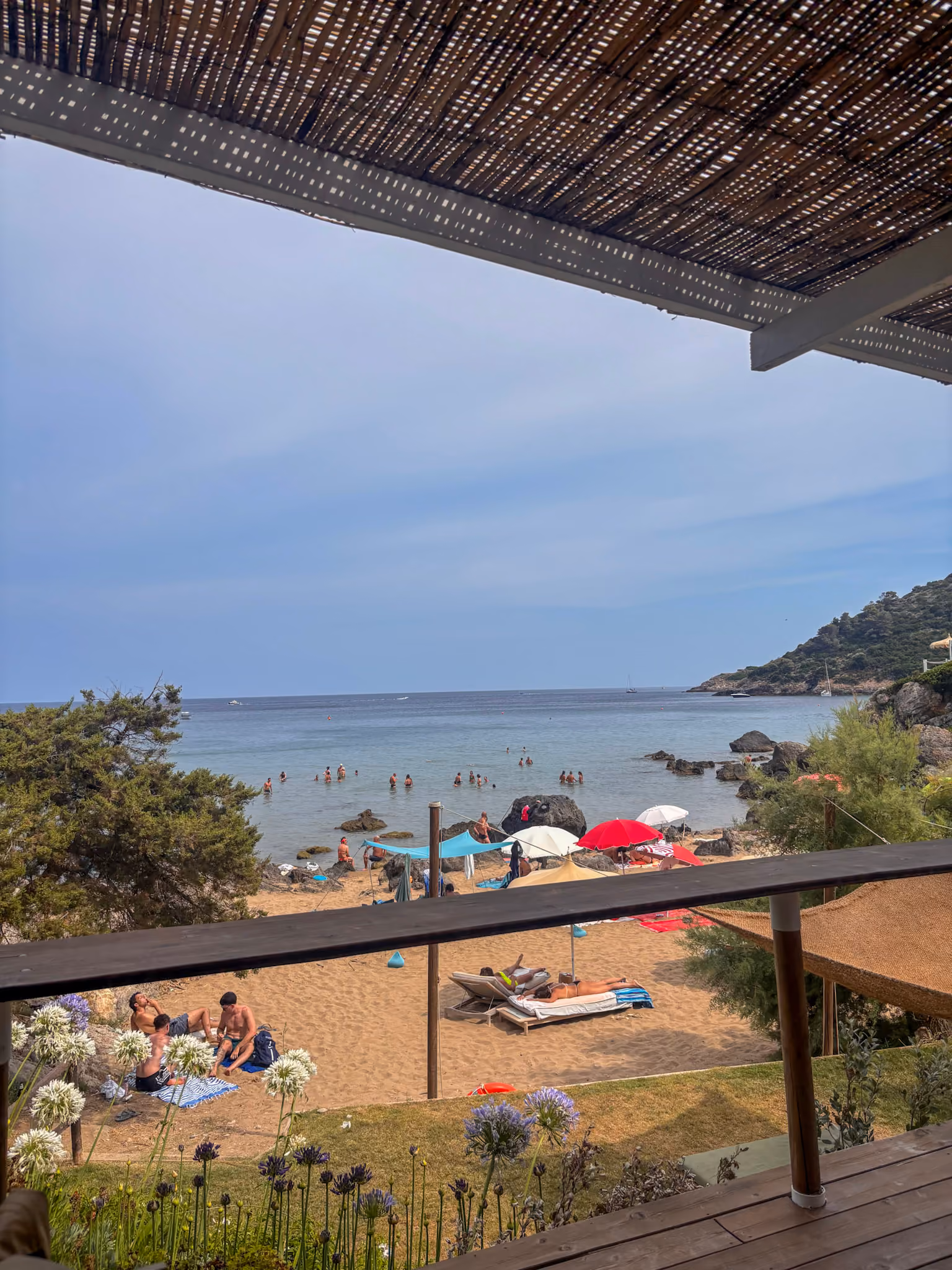 Strand mit Menschen, die im Sand liegen und im Meer schwimmen, Sicht von einer Terrasse mit Holzgeländer und einer überdachten Markise, blauer Himmel und hügelige Küstenlandschaft im Hintergrund.