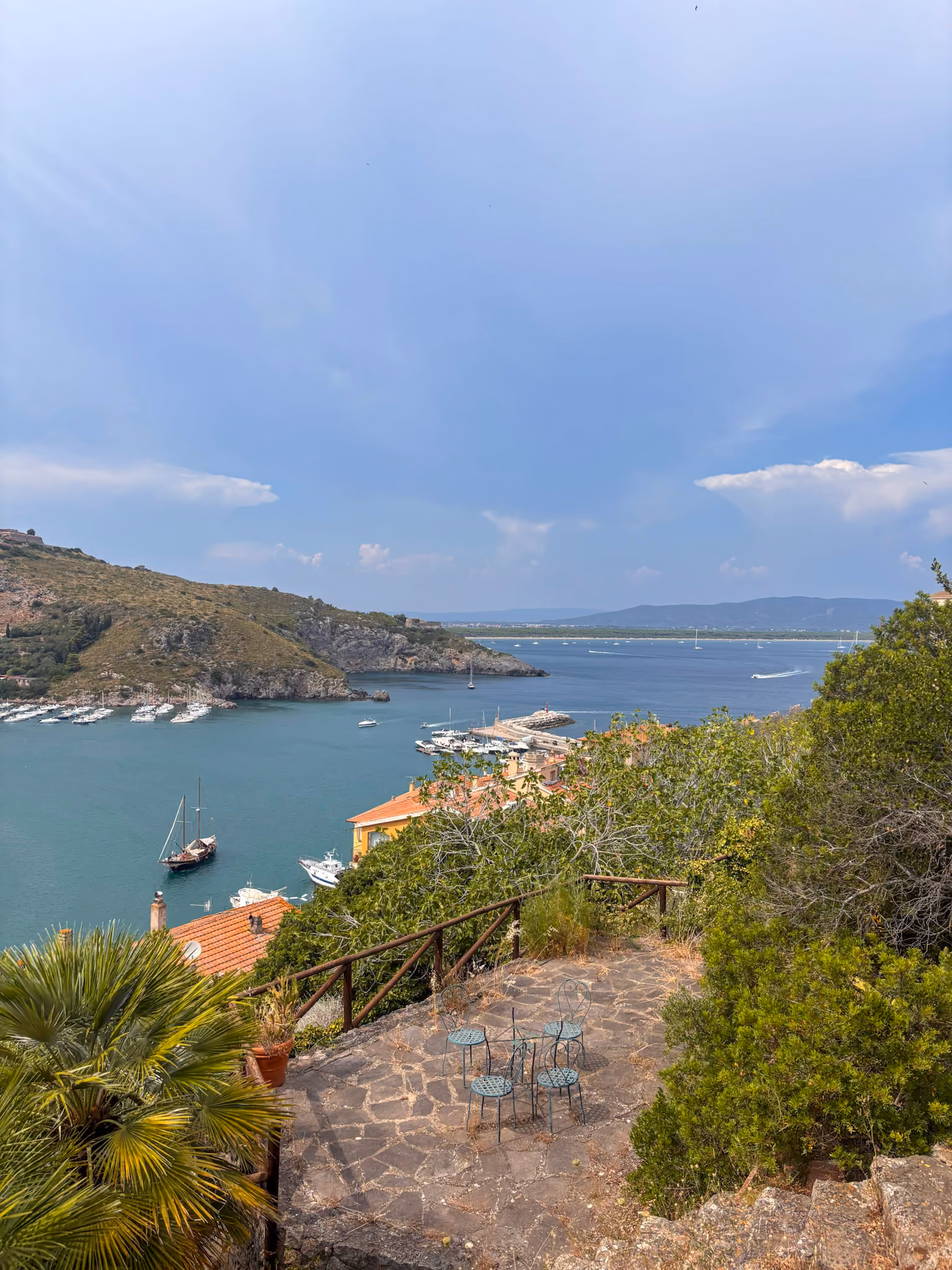 Blick von einer Terrasse mit Metallstühlen auf eine Bucht mit Booten, grünen Hügeln und blauem Himmel.