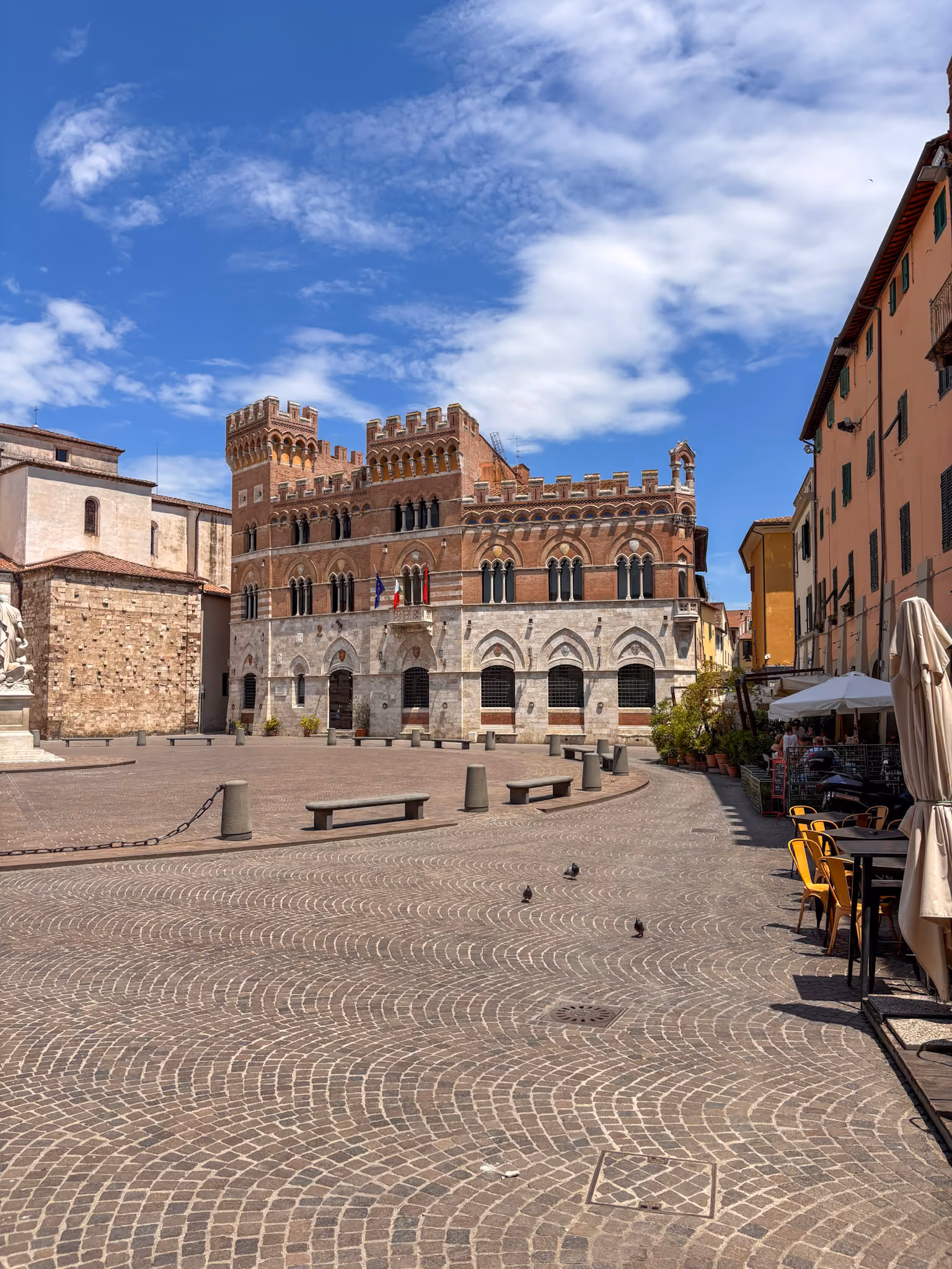 Historisches Gebäude mit Zinnen und mehreren Bögen auf einem gepflasterten Platz unter blauem Himmel, umgeben von weiteren Gebäuden und einem Straßencafé mit Tischen und Stühlen.