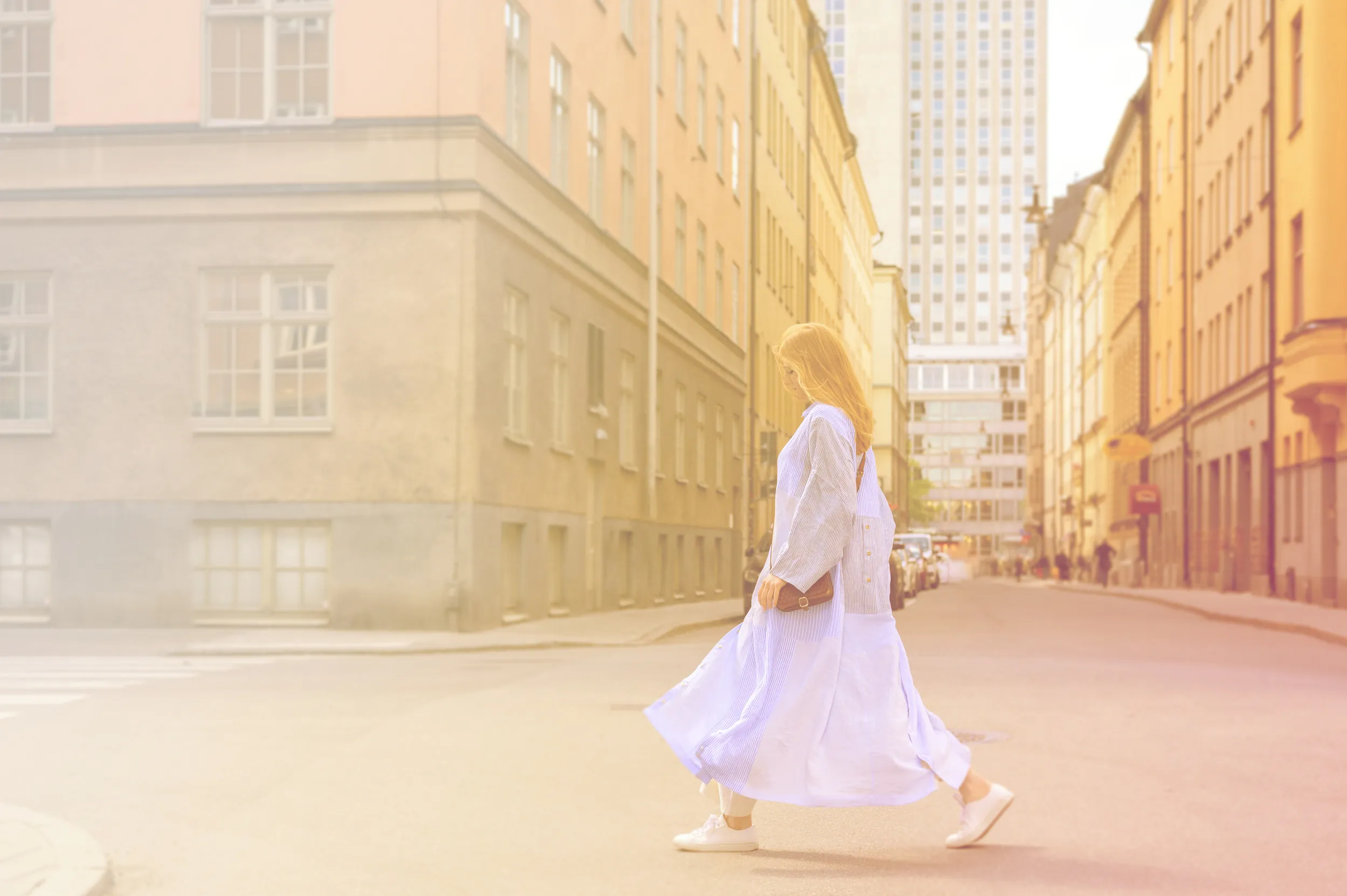 A woman walks in the south of Stockholm's inner city in a light blue long dress on the street. There are light leaks in the image as the sun is low before dusk.