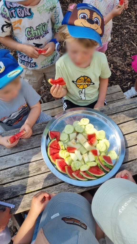 Elbzwerge-Kinder beim gemeinsamen Picknick am grauen Holztisch mit Wassermelone und Gurke an einem heißen Sommertag in Stellingen