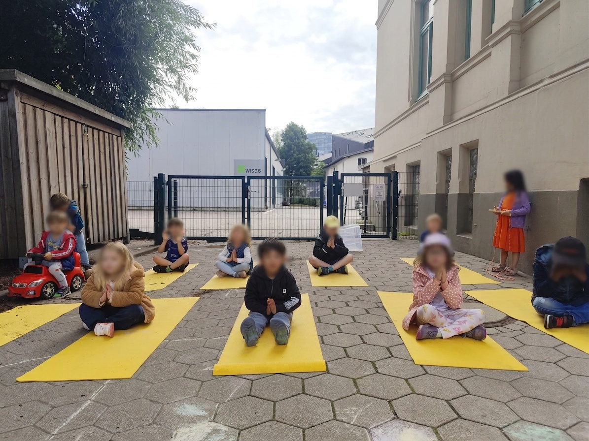 Kinder der elbzwerge Kita Harburg sitzen mit gefalteten Händen auf gelben Yogamatten im Innenhof