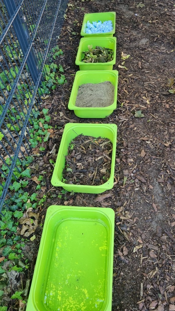 Fünf grüne Wannen mit Papier, Blättern, Sand, Mulch und Wasser im Garten der elbzwerge Kita Stellingen