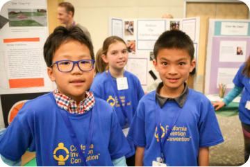 two young boys smiling while a young girl smiles in the background, all of them are wearing a blue CAIC shirt