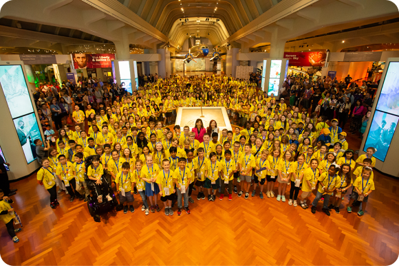 A high level picture of a huge room filled with hundreds of kids all wearing a yellow invention convention shirt
