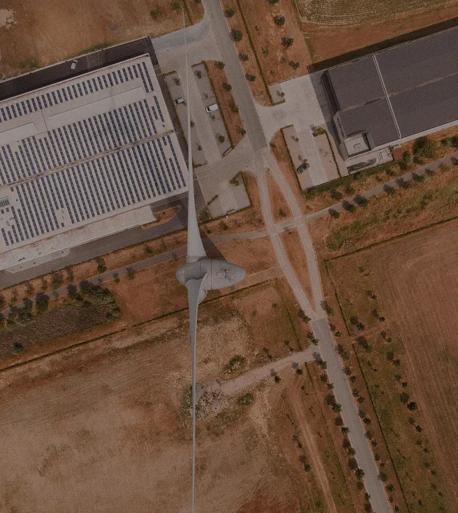 Aerial view of a large wind turbine next to industrial buildings with solar panels on the roof and surrounding dry farmland.