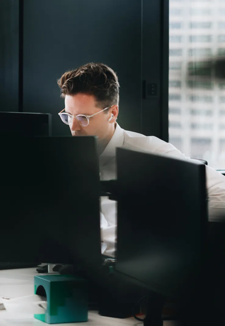 Man wearing glasses and white earbuds focused on work behind multiple computer monitors in an office.