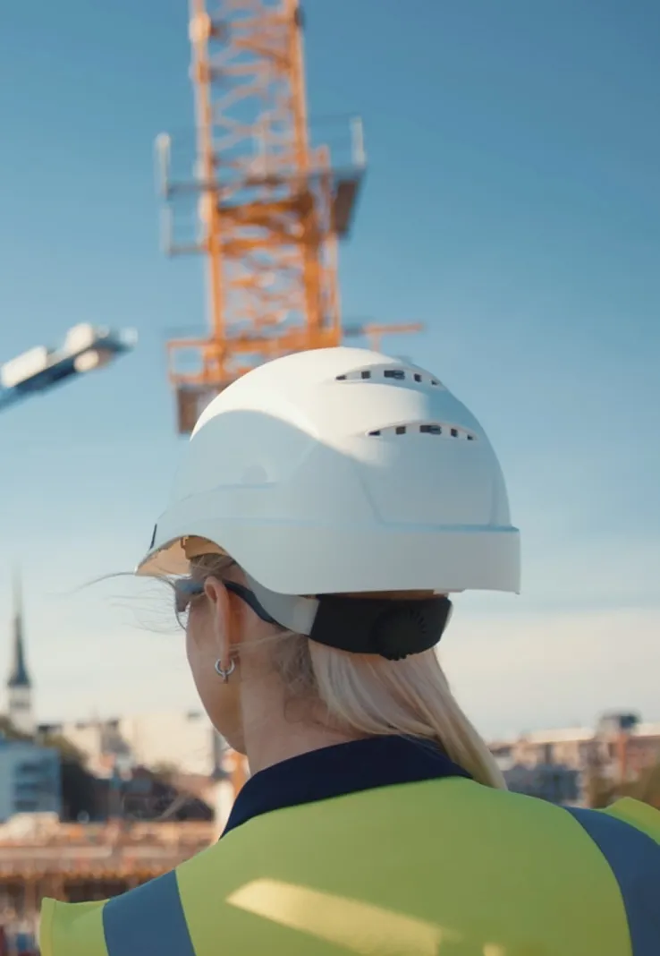 Rear view of a female construction worker with blonde hair wearing a white hard hat and yellow safety vest at a construction site with a crane in the background.
