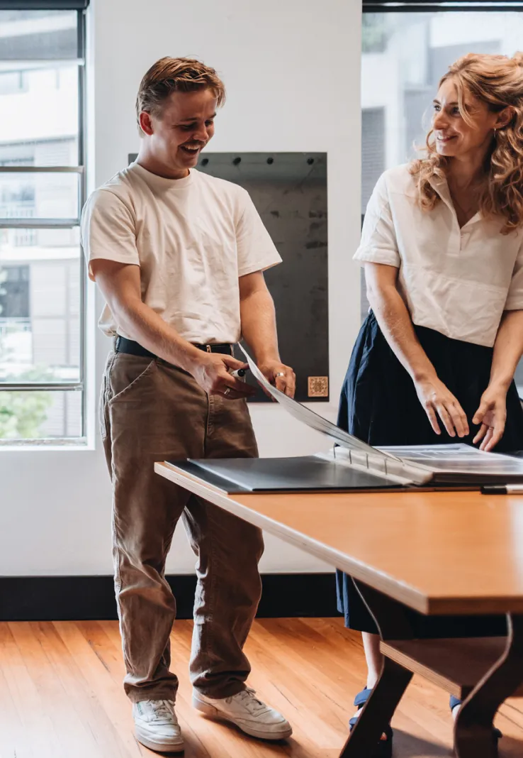Two young adults smiling and looking at documents on a table in a bright room with wooden floors.