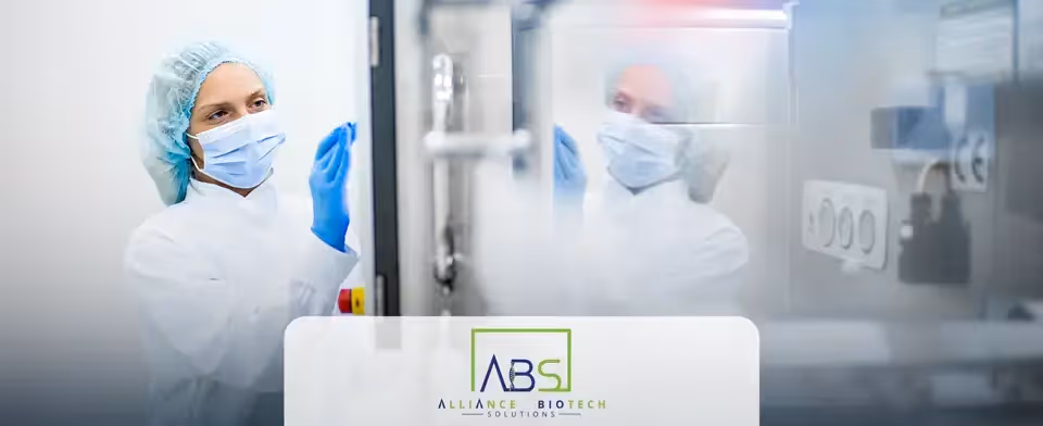 A woman wearing personal protective equipment assesses equipment in a laboratory setting. Her reflection appears in the glass. The Alliance Biotech Solutions logo is overlaid.
