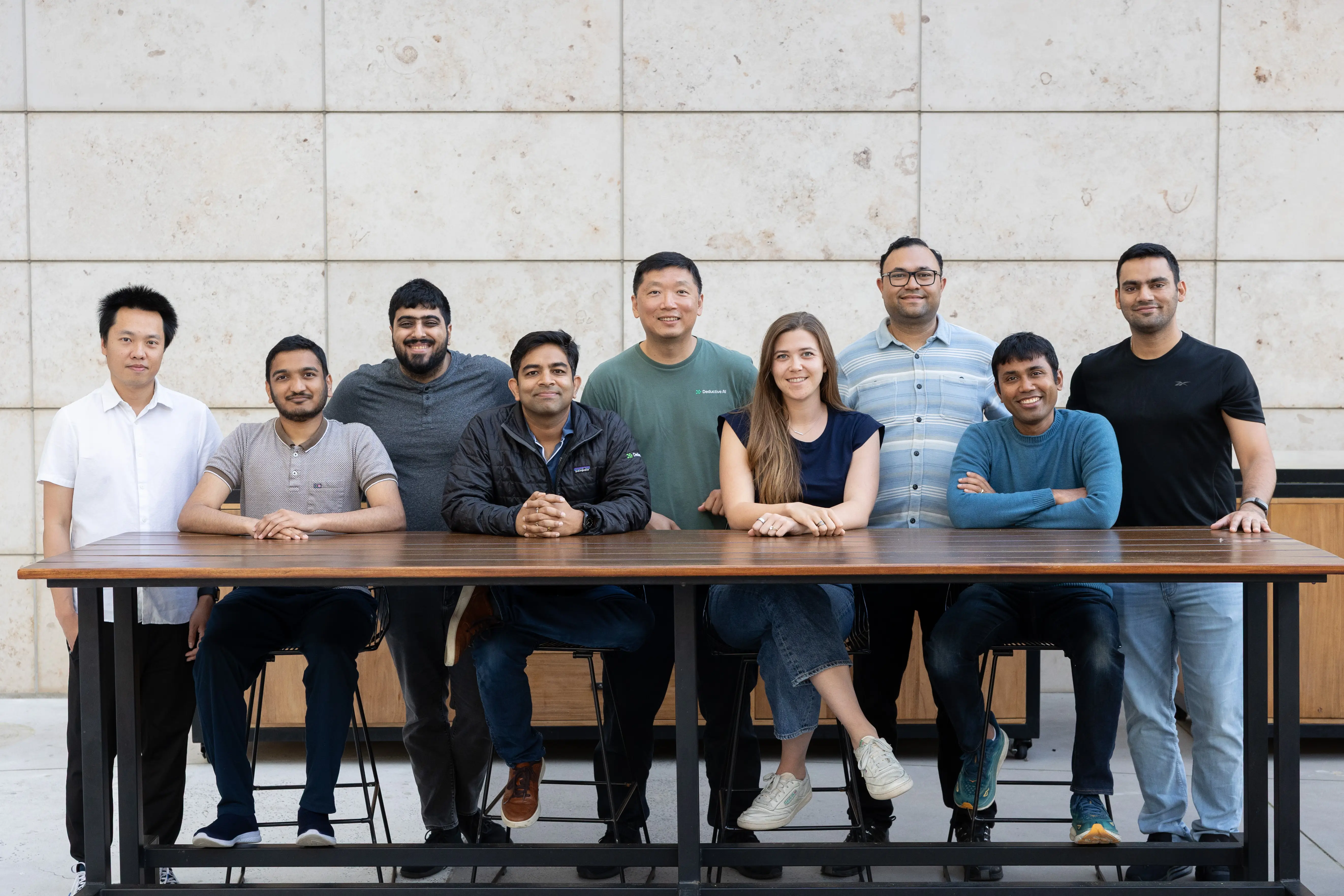 Deductive Team Picture where Group of nine diverse people smiling and posing behind a wooden table in an indoor setting with a tiled wall background.
