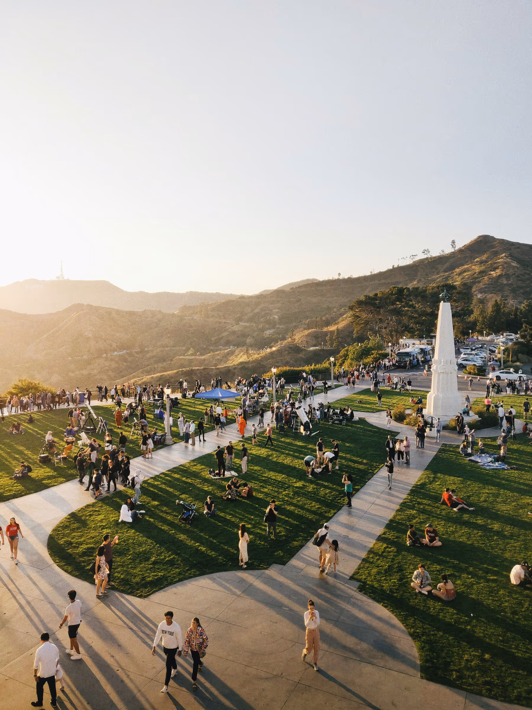 Crowd of people gathered on green lawns with walkways and a white obelisk monument in a park, with hills in the background under clear sky.