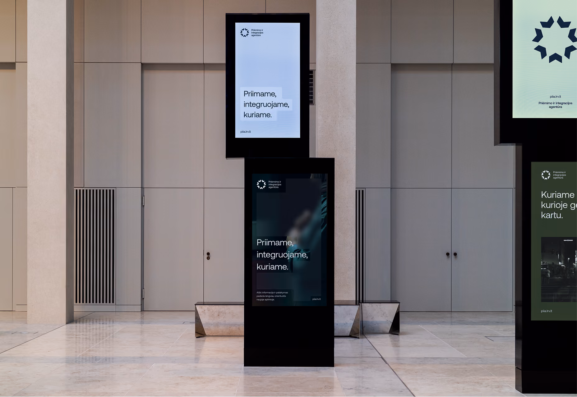 Two digital info boards in a building lobby with Lithuanian text about integration agency.