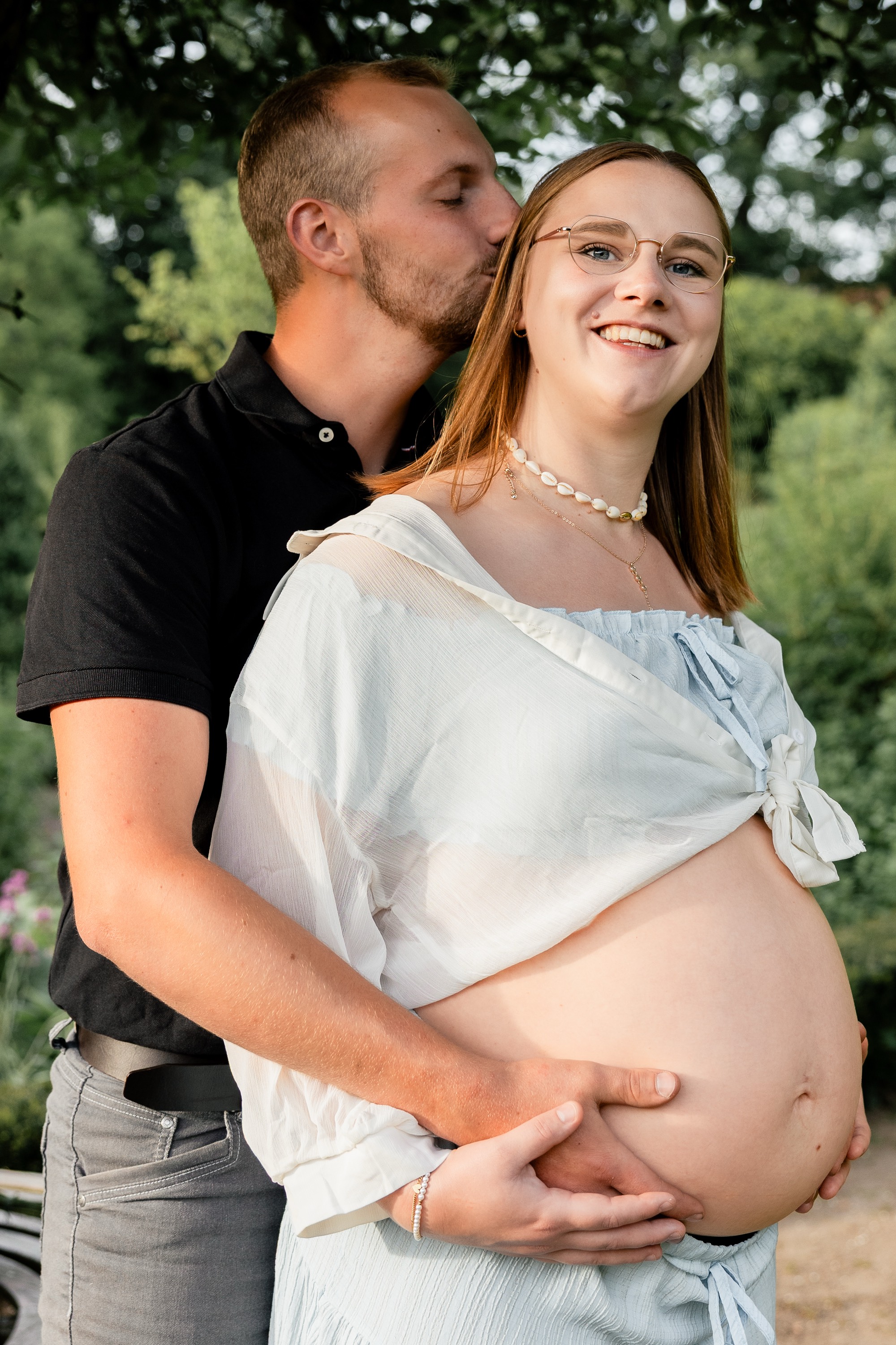 Werdender Vater küsst seine schwangere Partnerin an der Schläfe, beide Hände auf dem Babybauch