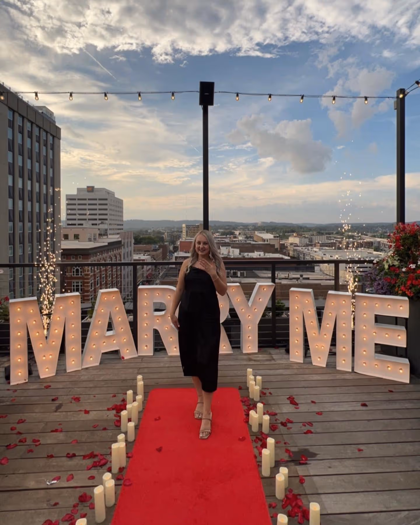 Woman in a black dress standing on a red carpet surrounded by lit candles and rose petals with illuminated letters spelling 'MARRY ME' behind her on a rooftop at sunset.