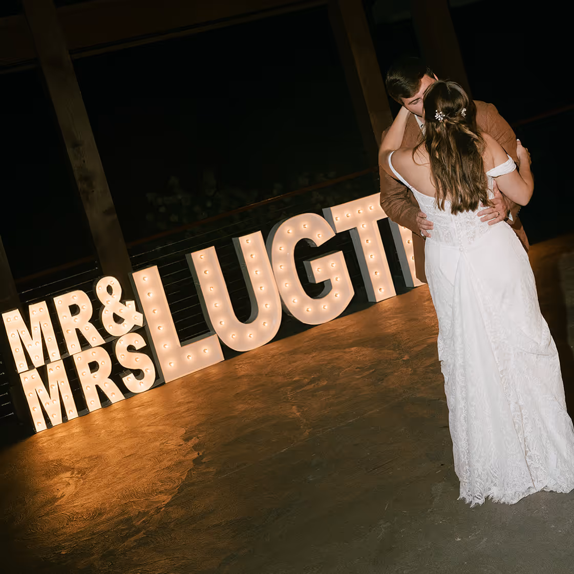 Bride and groom sharing a kiss on the dance floor next to illuminated letters spelling 'MR & MRS LUGHTI'.