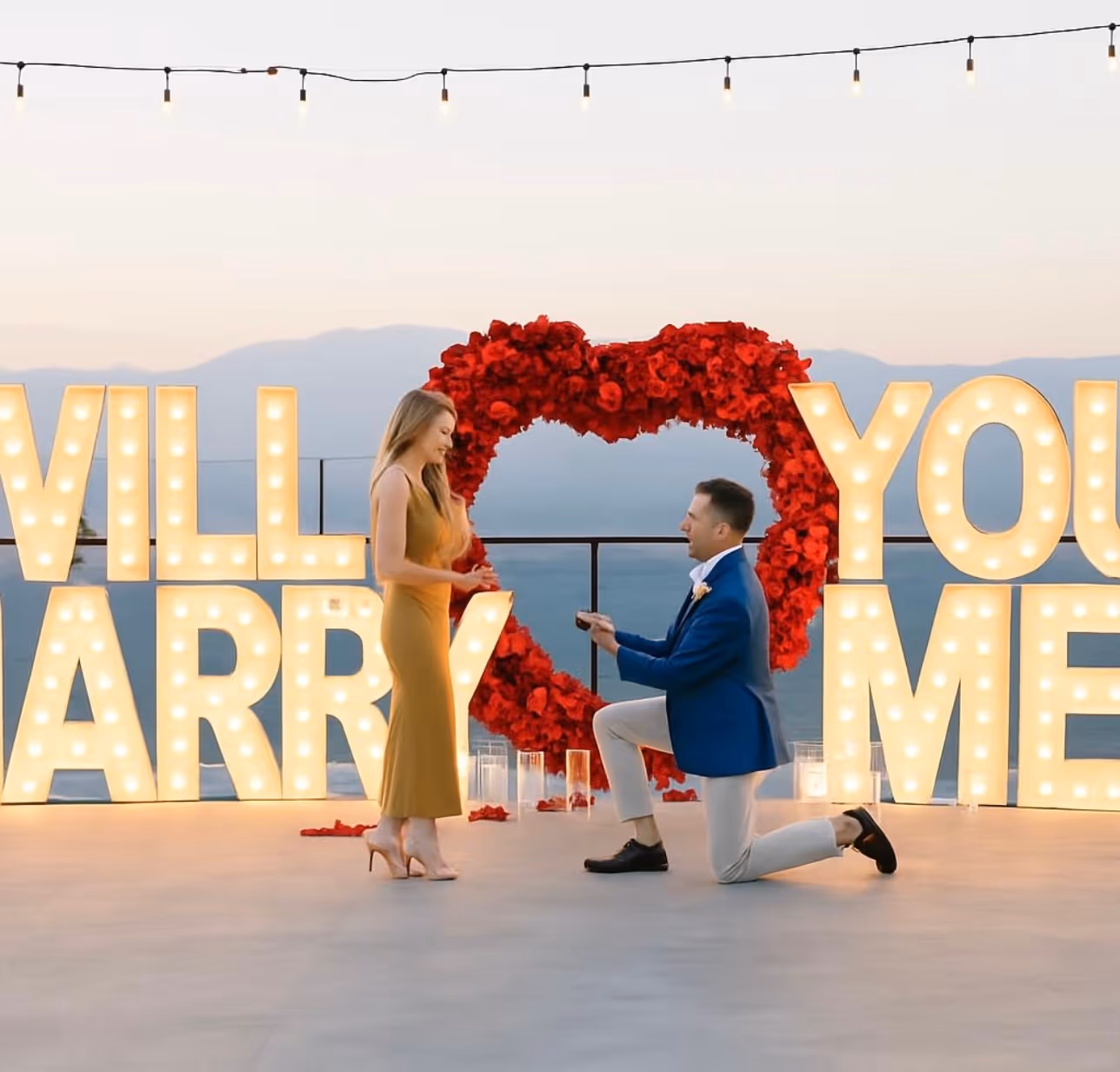 Man in blue blazer kneeling and proposing to woman in mustard dress in front of illuminated 'Will You Marry Me' sign with red rose heart backdrop.
