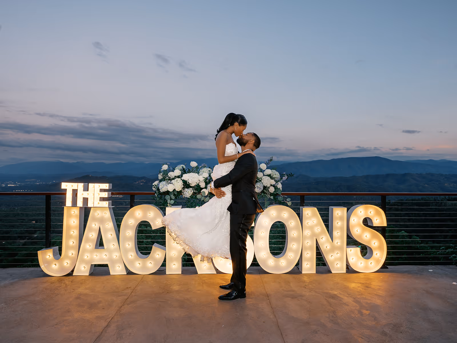 Bride and groom embracing in front of large lighted letters spelling THE JACKSONS with mountains in the background at dusk.
