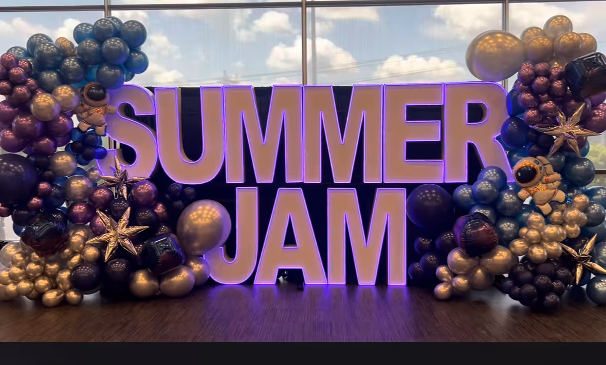 Large illuminated letters spelling 'SUMMER JAM' surrounded by clusters of purple, black, and gold balloons with space-themed decorations including astronauts and stars.