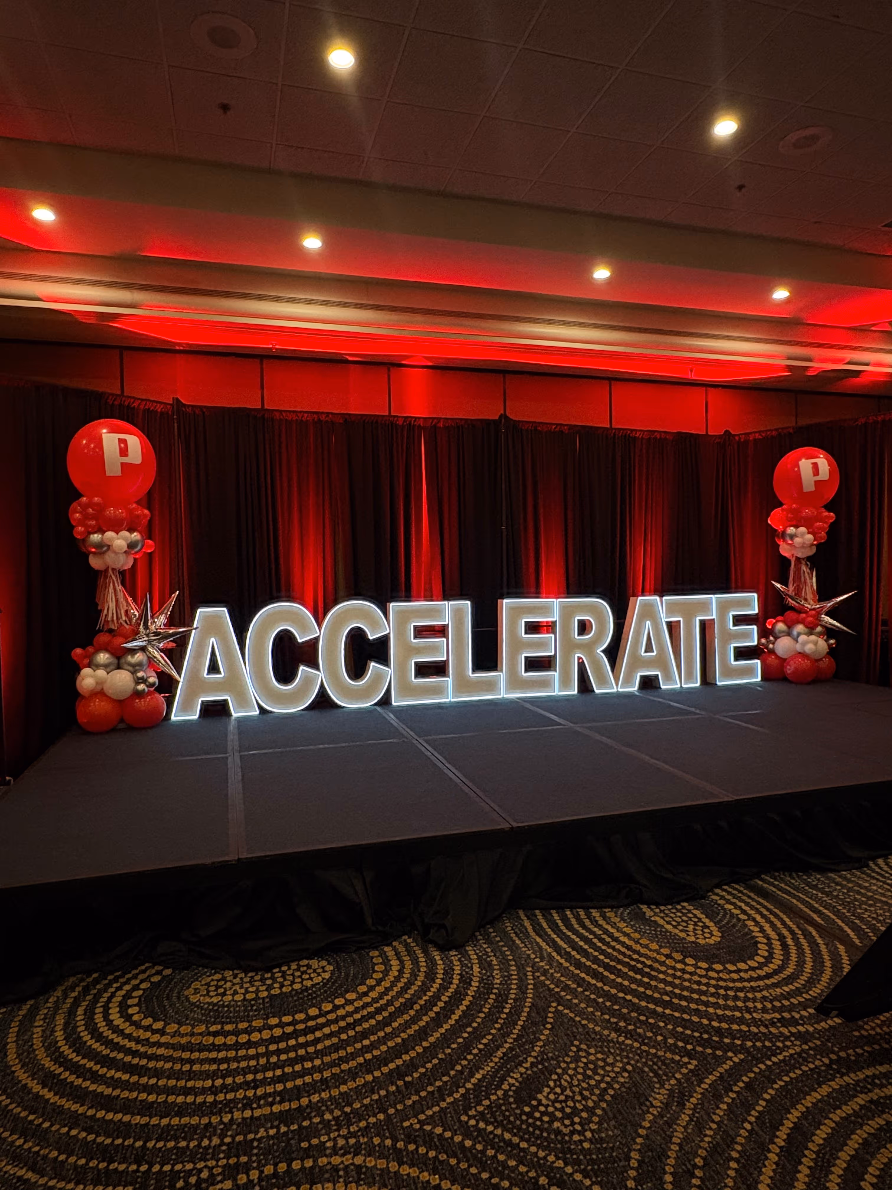 Stage with large illuminated letters spelling ACCELERATE, flanked by red and white balloon columns with silver stars.