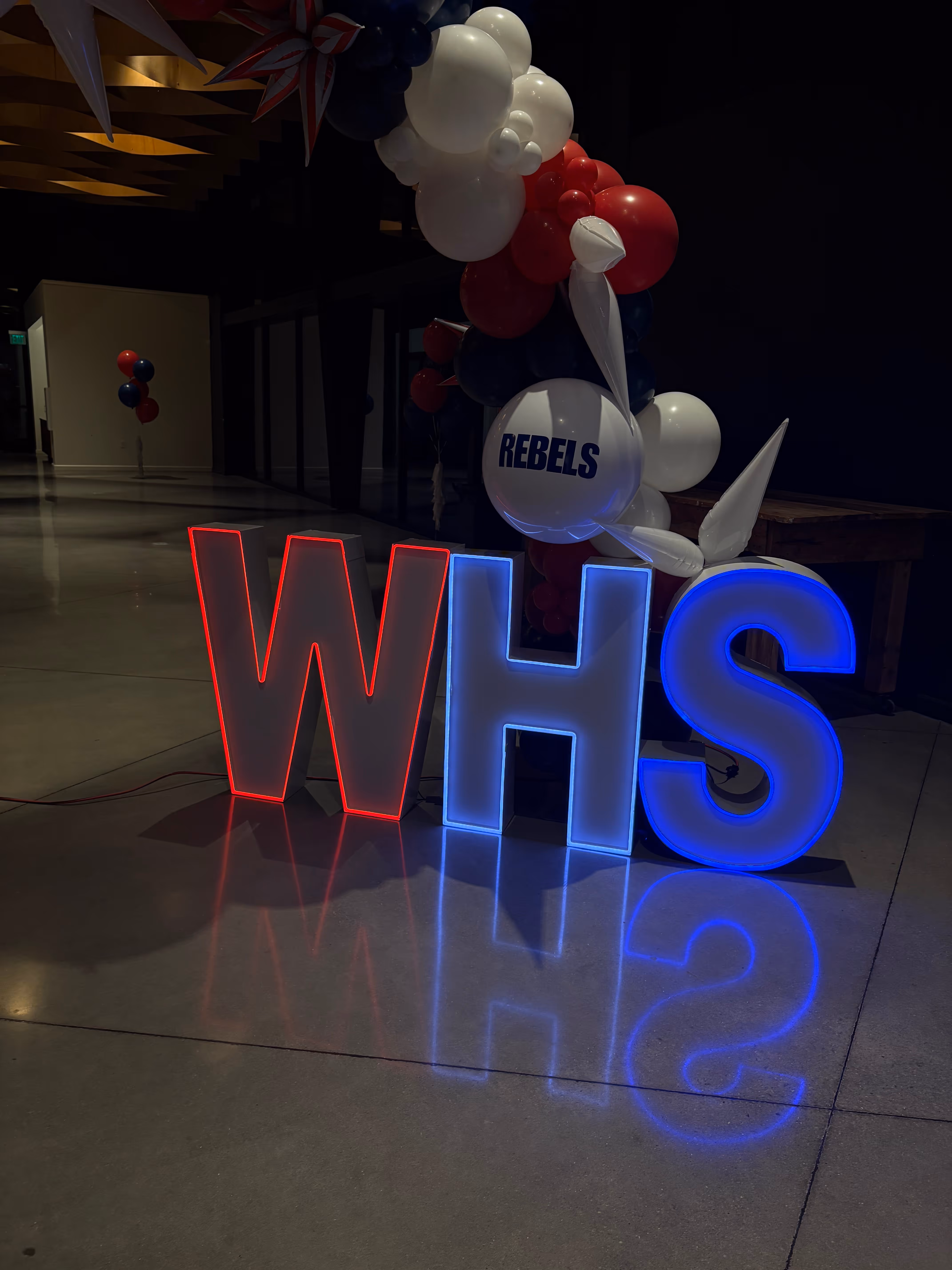 Large illuminated letters W, H, S in red and blue light with balloons spelling REBELS in a dimly lit room.