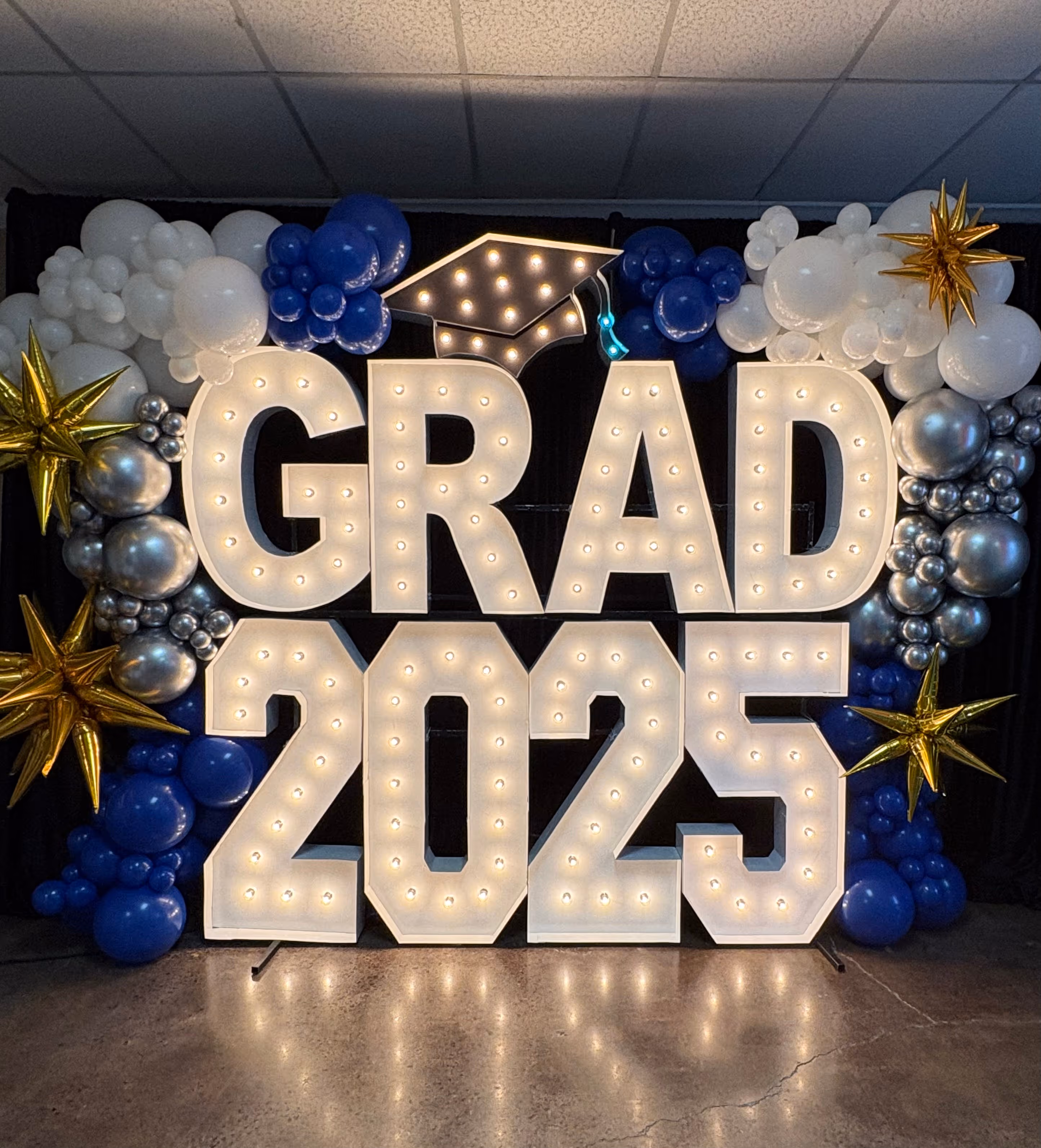 Illuminated marquee letters spelling GRAD 2025 surrounded by blue, white, silver balloons and gold star-shaped decorations with a lit graduation cap on top.
