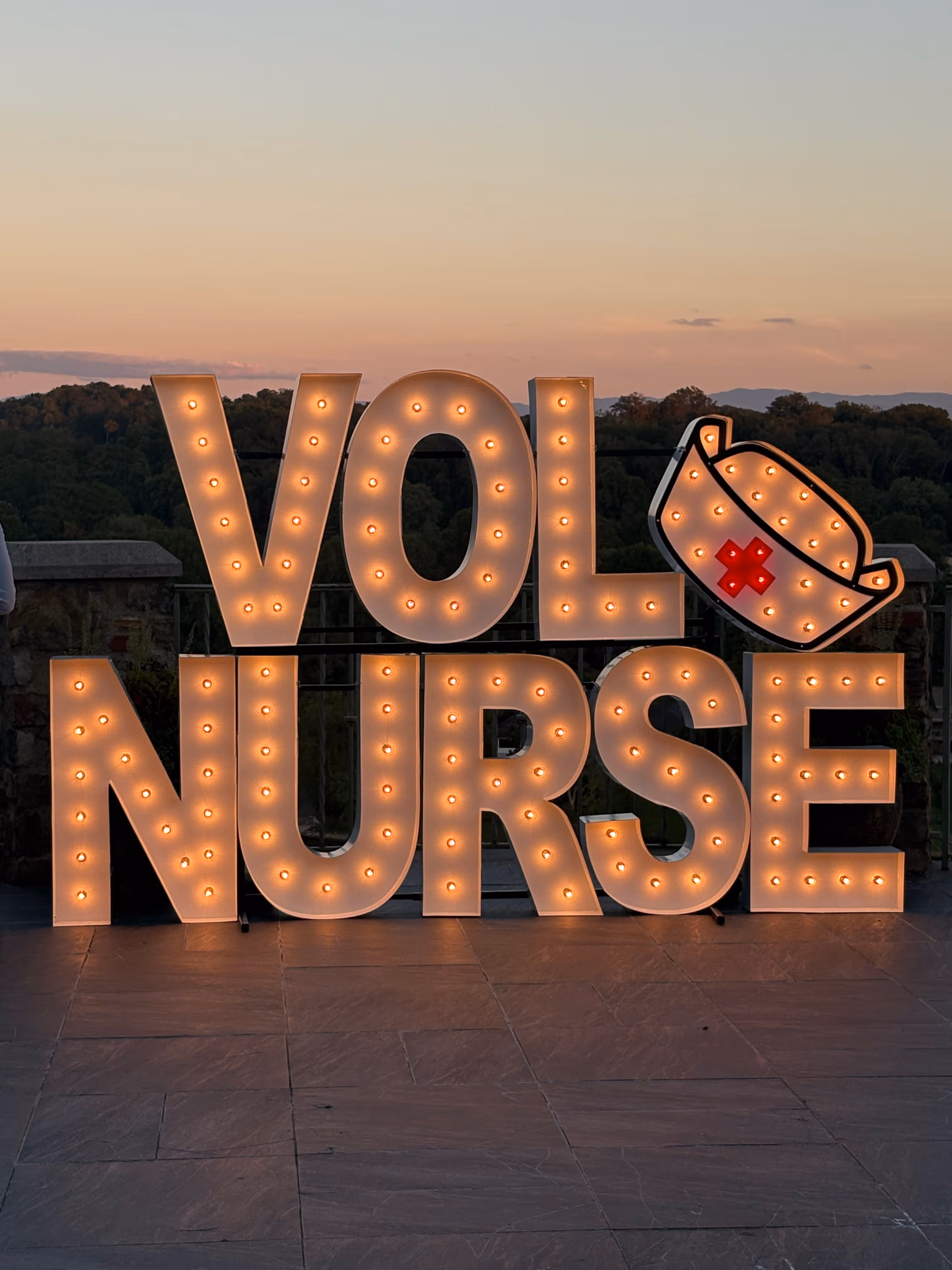 Illuminated sign reading VOL NURSE with a nurse's cap featuring a red cross on the letter E, set outdoors at sunset.