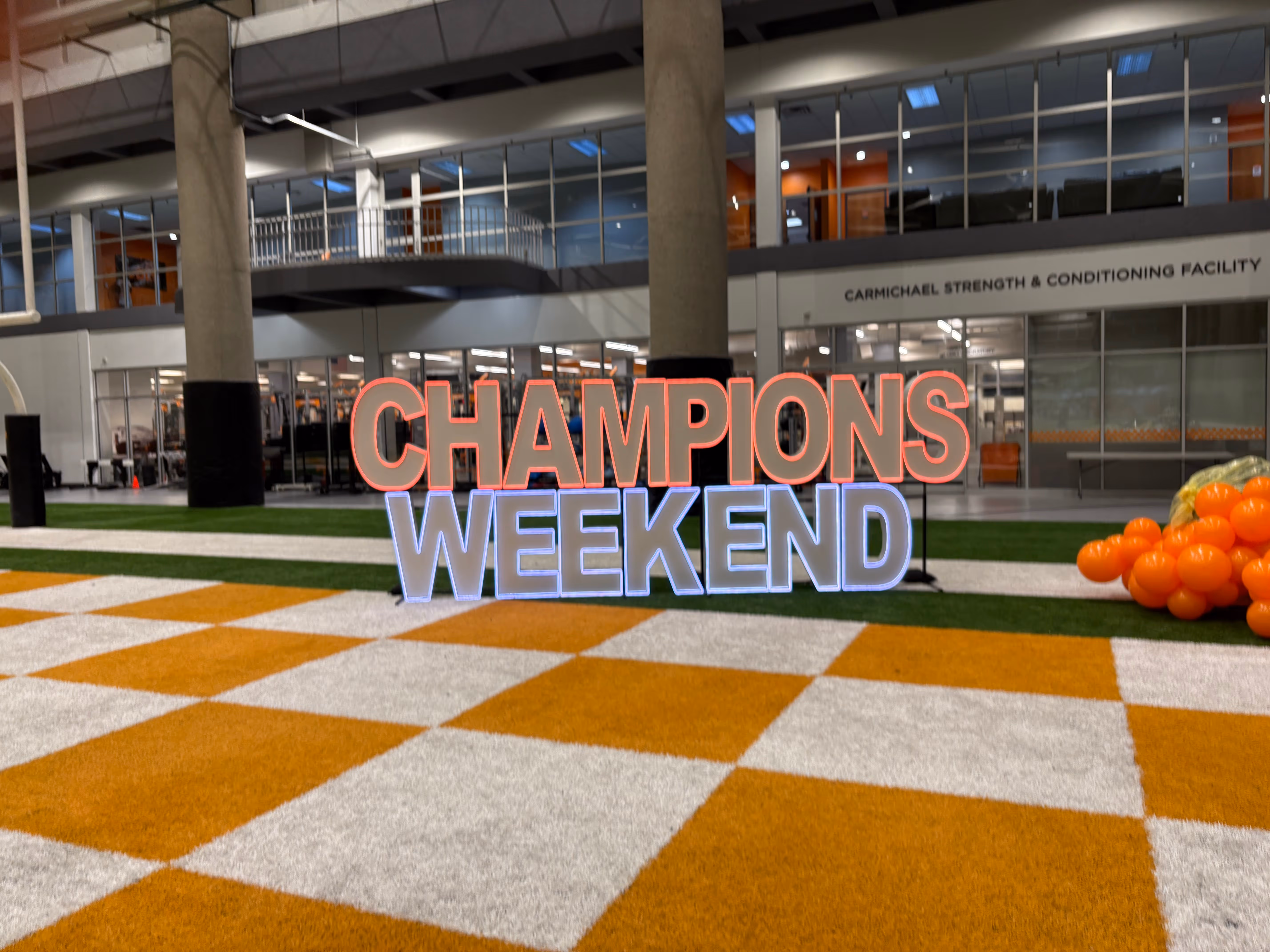 Indoor sports facility with a neon sign reading 'CHAMPIONS WEEKEND' on an orange and white checkered floor next to a bunch of orange balloons.