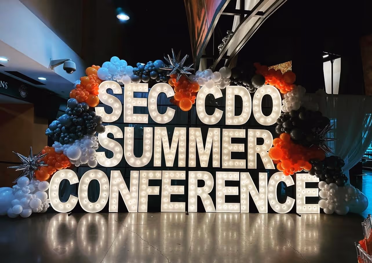 Illuminated letters spelling 'SEC CDO SUMMER CONFERENCE' surrounded by clusters of black, white, and orange balloons with silver star-shaped balloons.