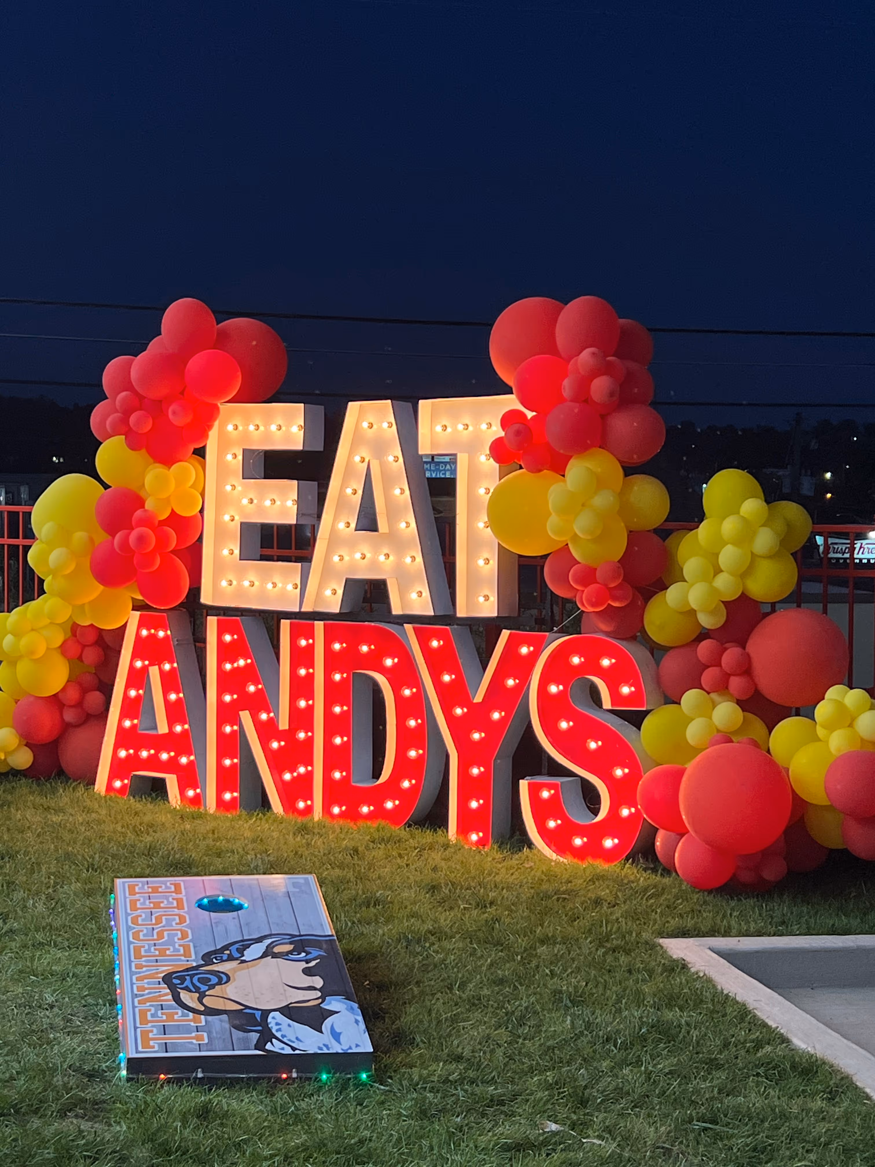 Illuminated marquee letters spelling 'EAT ANDYS' decorated with red and yellow balloons, displayed outdoors at night with a cornhole board on grass in front.