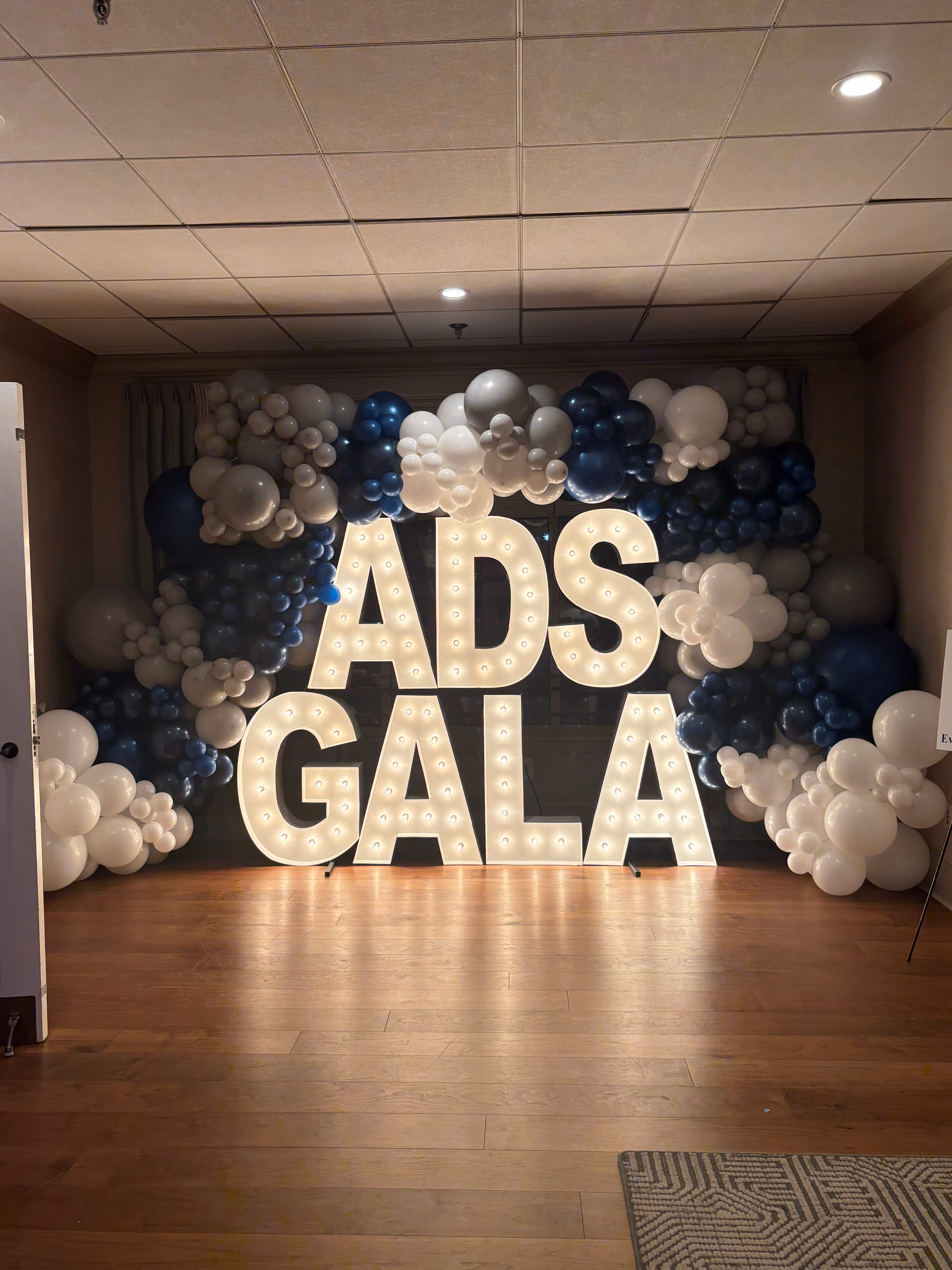 Illuminated white letters spelling 'ADS GALA' surrounded by a large balloon arrangement in white and dark blue colors.