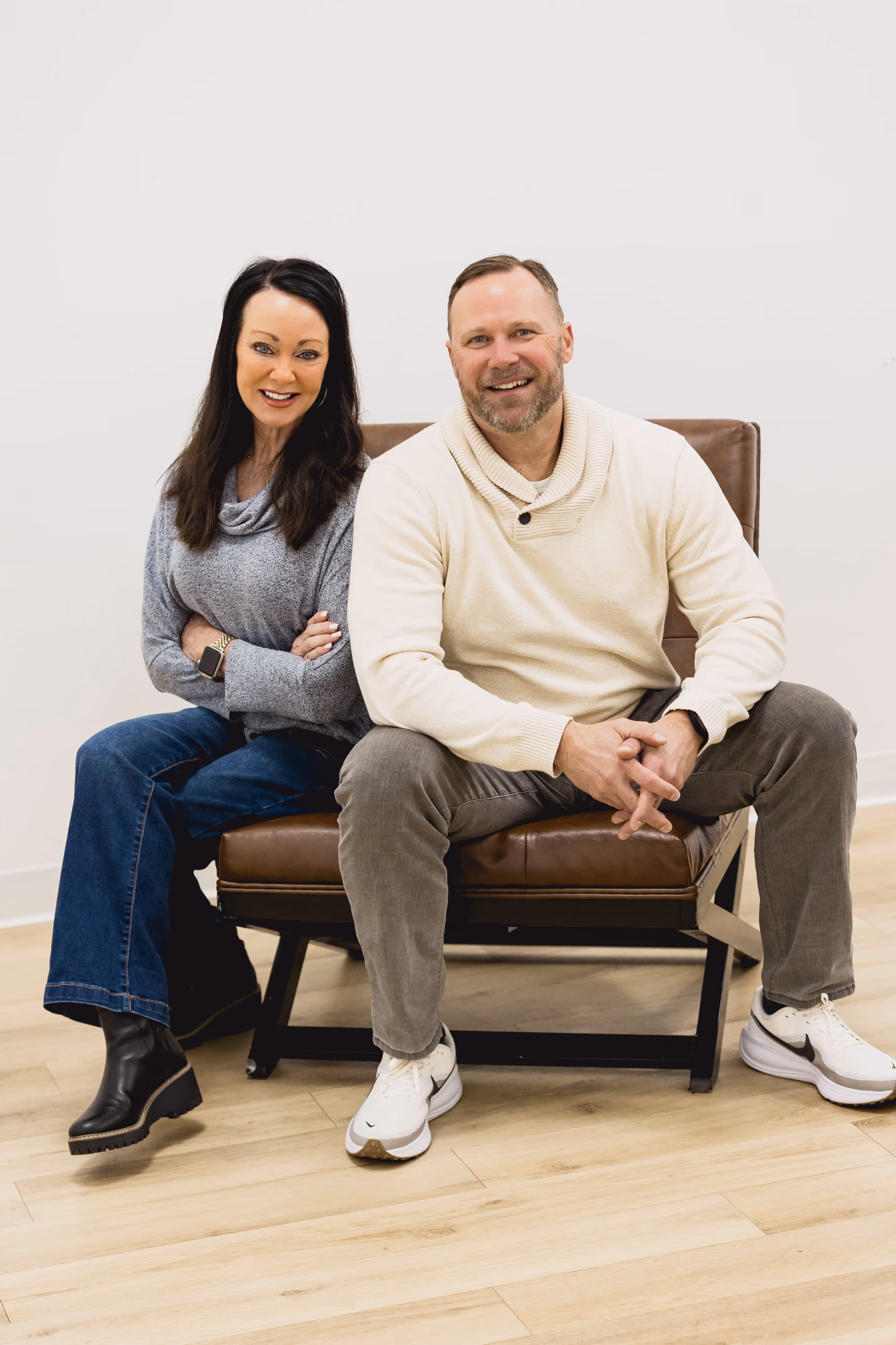A smiling man and woman sitting side by side on a brown leather chair against a plain white wall.