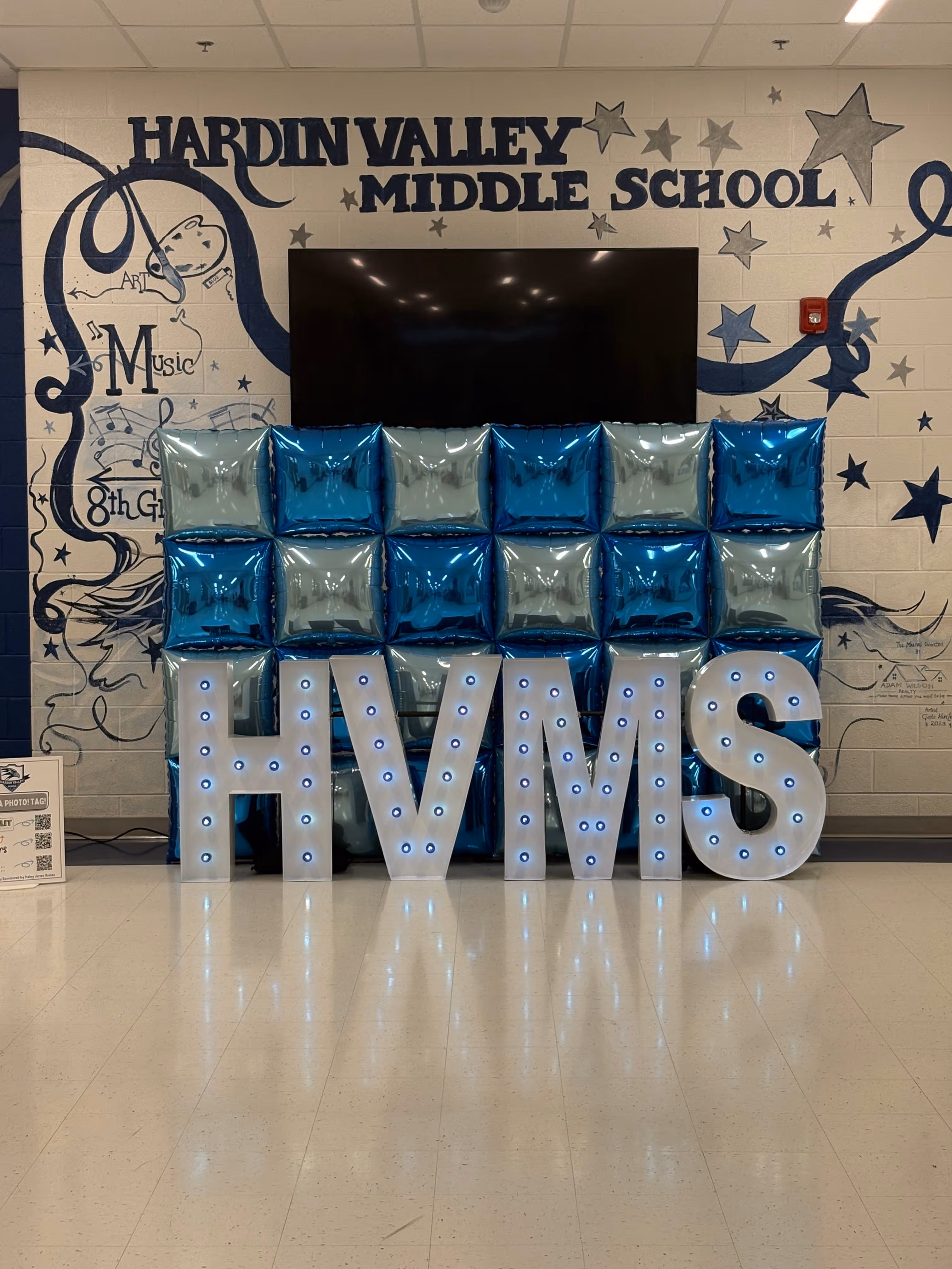 Lit letters spelling HVMS in front of silver and blue balloons at Hardin Valley Middle School with mural stars and music notes on the wall.