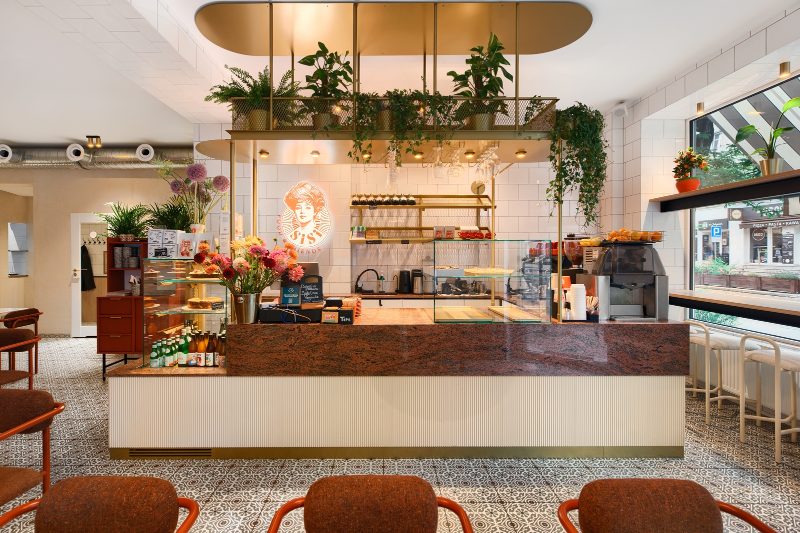 Modern cafe interior with patterned tile floor, a counter with glass display cases, flowers, hanging plants, and seating with brown cushioned chairs.