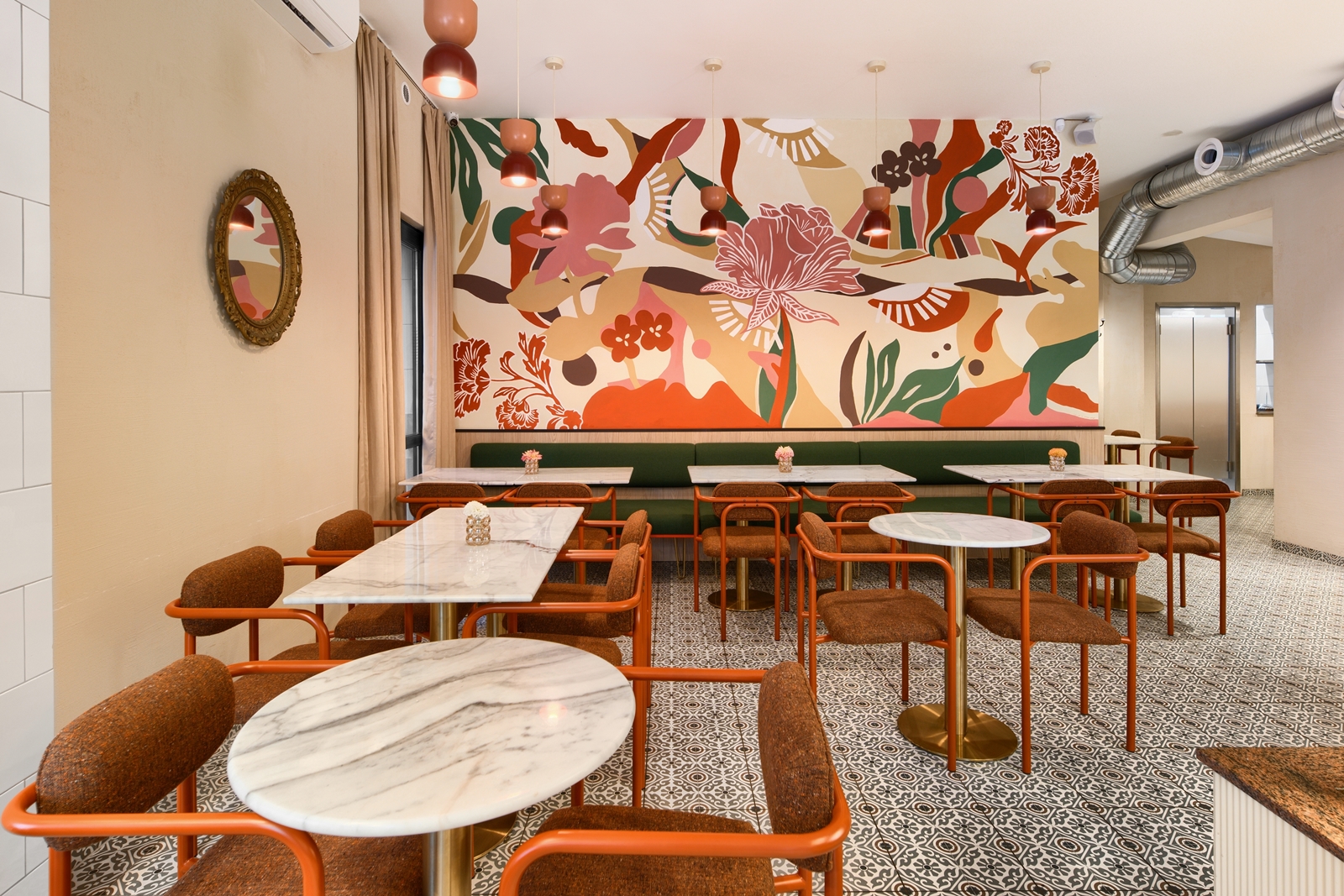 Modern café interior with marble tables, rust-colored chairs, a green bench, patterned tile floor, and a colorful floral mural on the back wall.