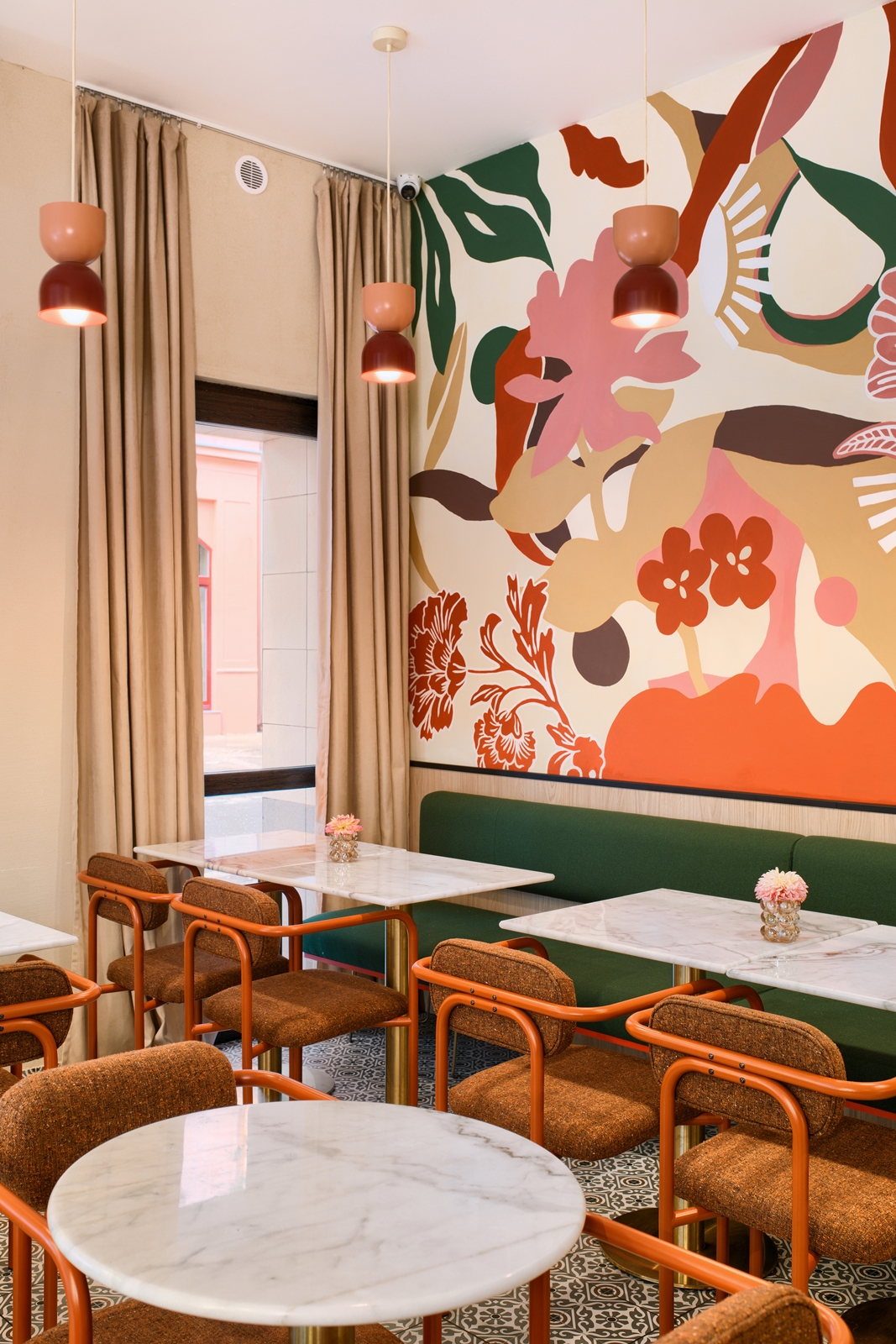 Modern cafe interior with marble tables, brown cushioned chairs with orange frames, green bench seating, hanging pendant lights, and a colorful abstract floral mural on the wall.