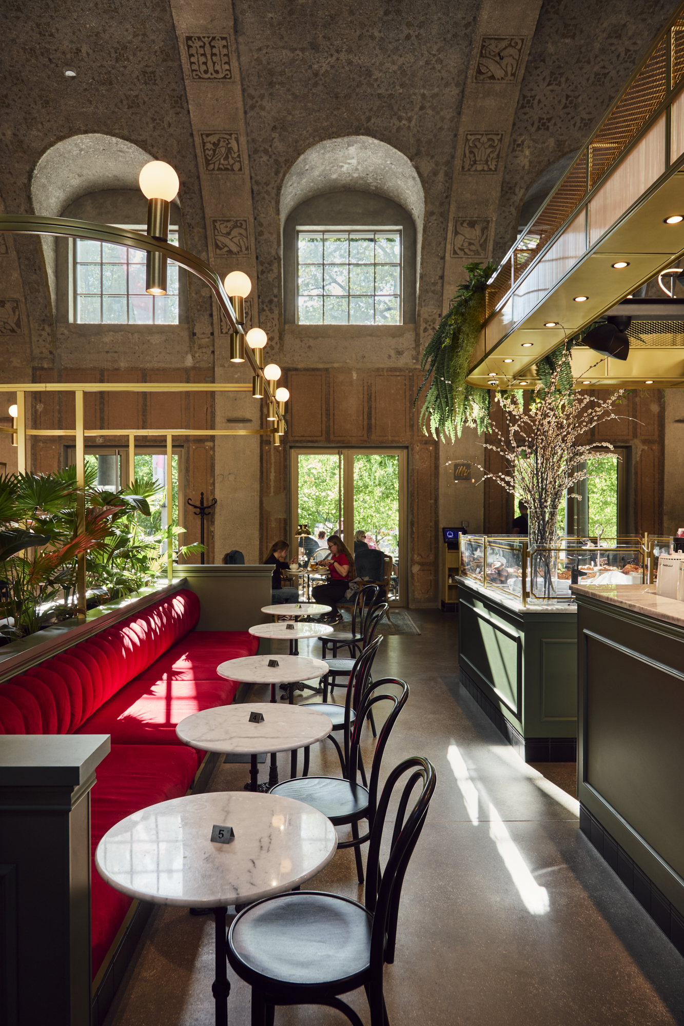 Interior of a café featuring a long red velvet bench, marble round tables with black chairs, large arched windows, and a display counter with plants and lighting.