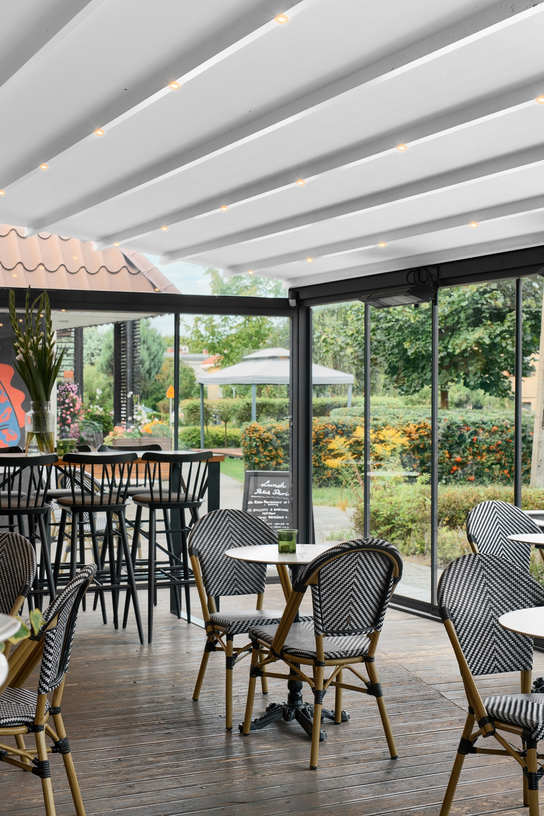 Indoor café seating with black and white woven chairs, wooden floor, and glass walls overlooking a garden with bushes and flowers.