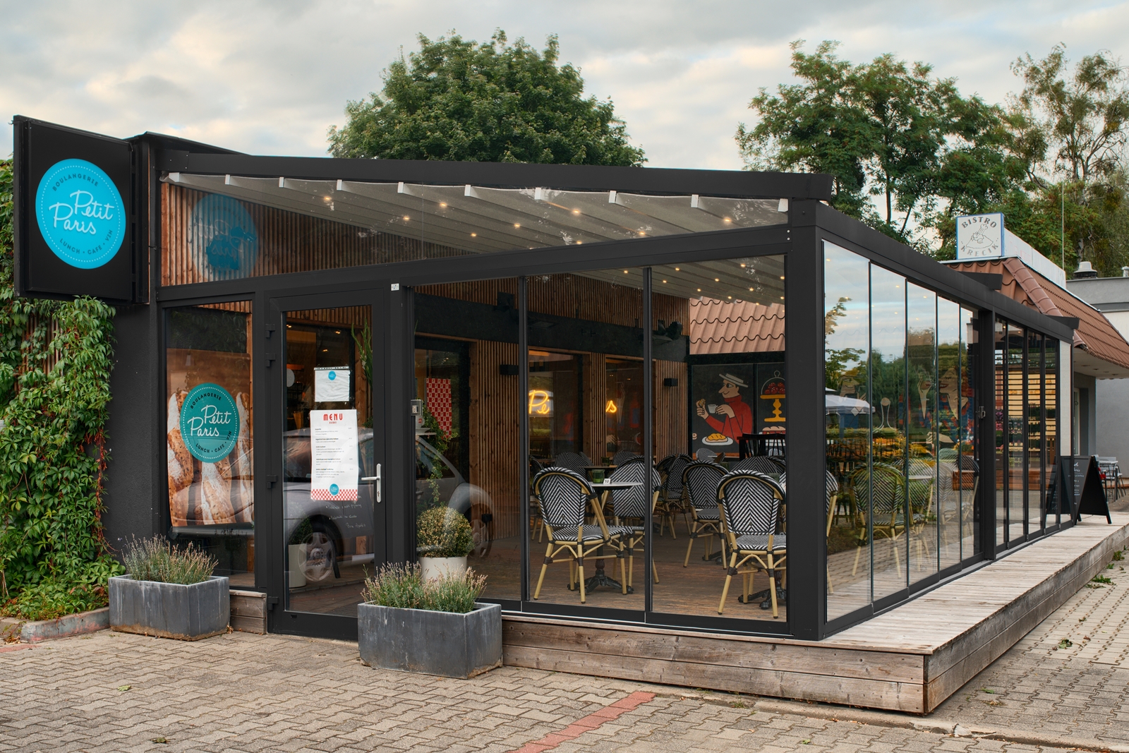Modern glass-enclosed café with black-framed structure, outdoor planters, and black-and-white patterned chairs inside.
