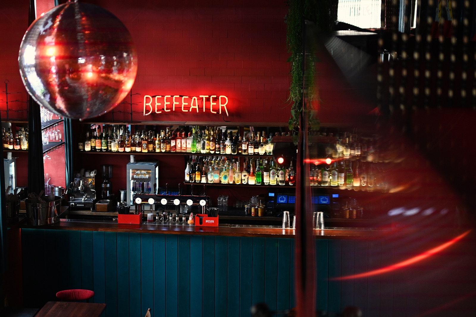 Bar interior with a teal counter, shelves stocked with liquor bottles, a disco ball hanging from the ceiling, and a red neon sign reading 'BEEFEATER'.
