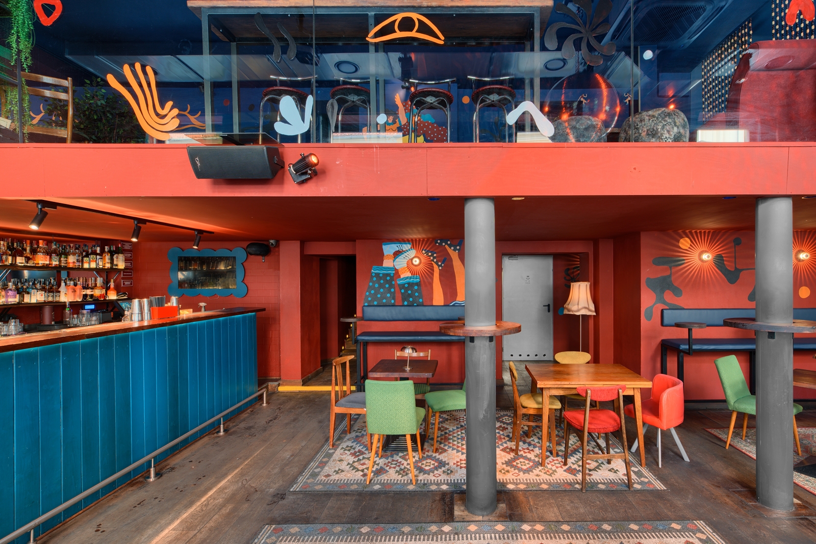 Cozy modern café interior with orange-framed brown chairs, white marble tables, green cushioned bench seating, and a colorful floral mural on the wall.