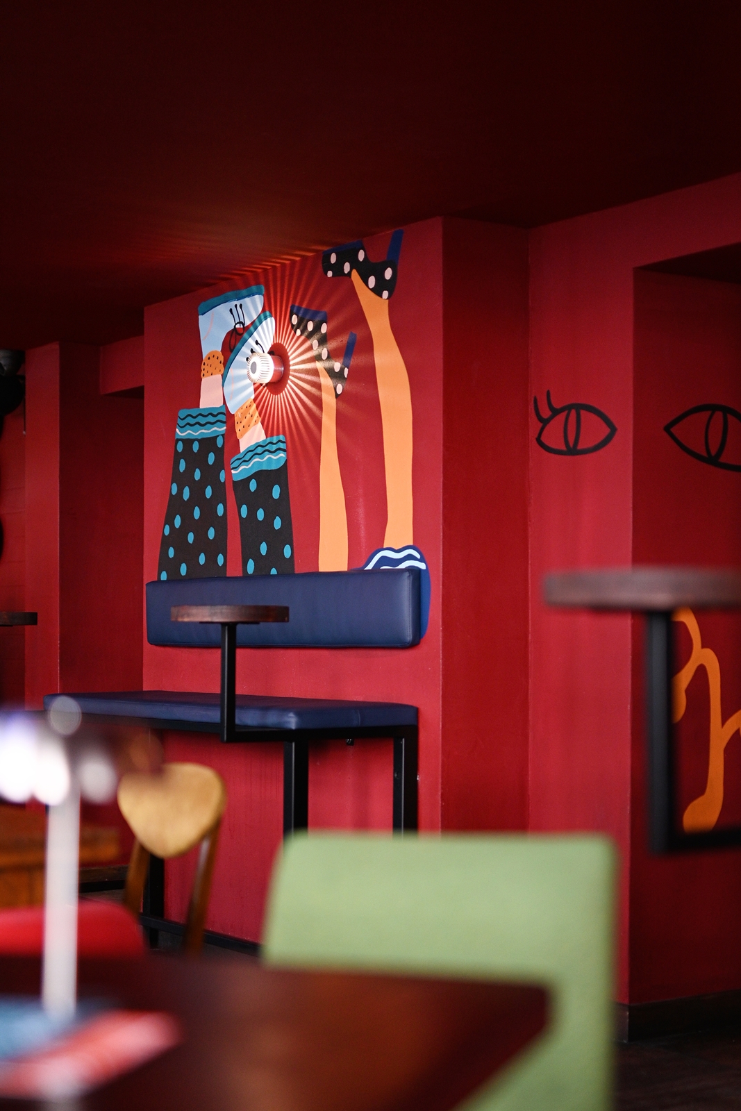 Red-walled room with a mural of colorful legs wearing patterned socks and shoes, and seating with a blue bench and wooden tables.
