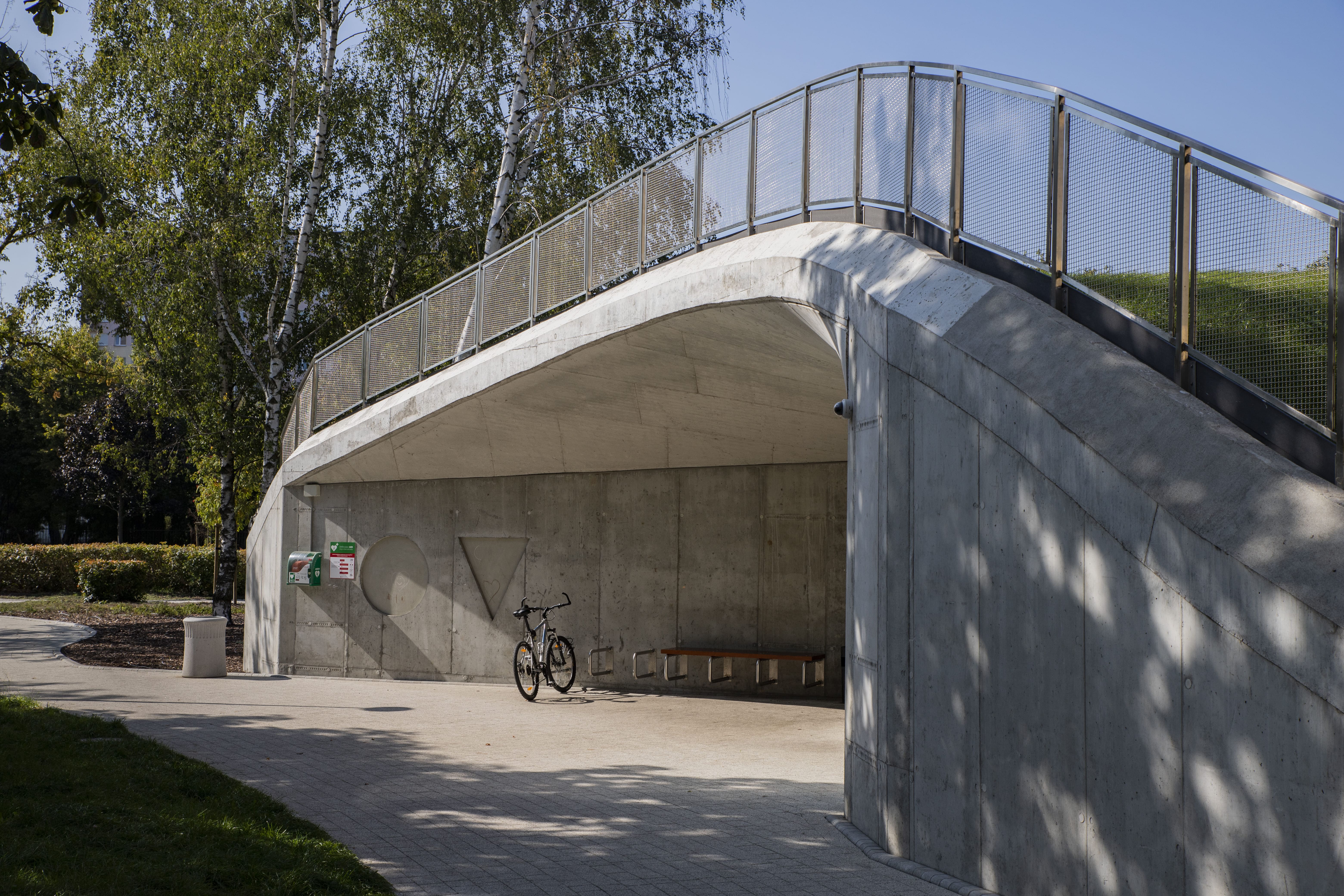 Concrete pedestrian bridge with metal railings over a pathway, a bicycle parked beneath it, surrounded by trees and greenery.
