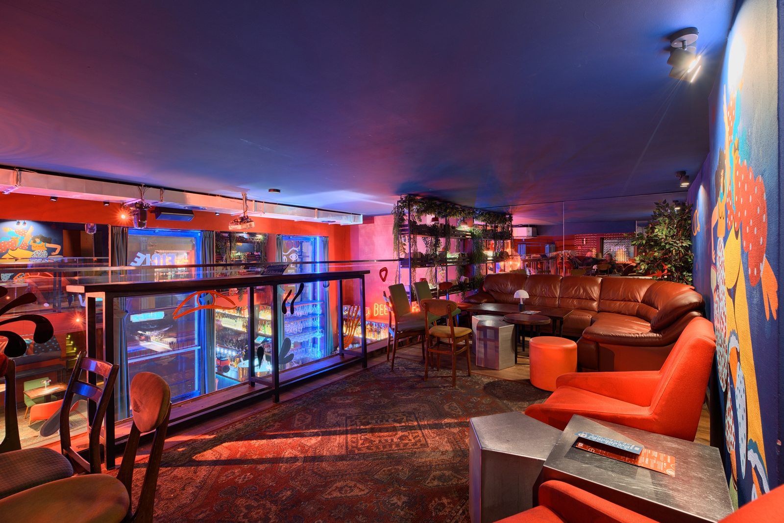 Cozy lounge area with a red sectional sofa, wooden chairs, a blue ottoman, a patterned rug, blue walls with colorful mural art, and hanging greenery.